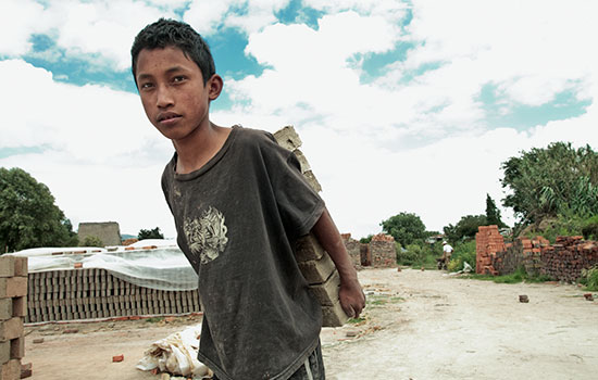A boy in a gray t-shirt carries a stack of bricks on his back