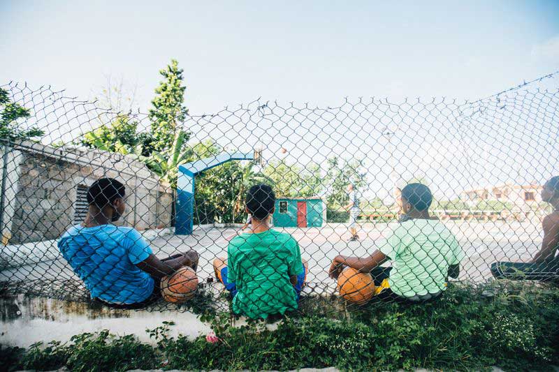 Four boys take a break from playing and sit against a fence with their basketballs