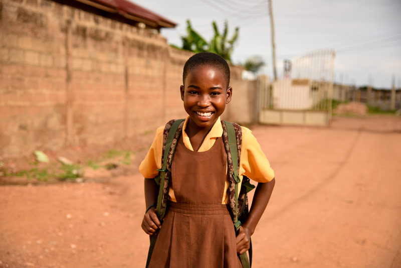 A smiling girl wears a backpack while walking down a dirt road
