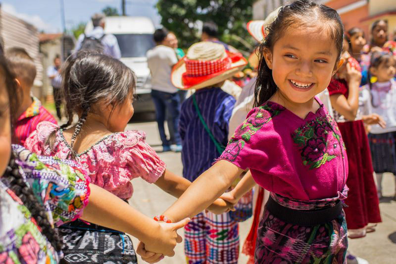 A group of little girls in traditional dresses hold hands as they spin around and smile