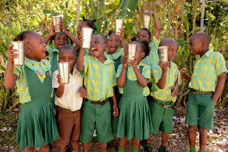 A group of children in a rural community smile and hold up glasses of clean water
