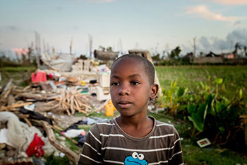 a Haitian child stands in front of a home destroyed by Hurricane Matthew