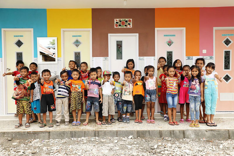 A group of children stand in front of a colorful wall and give a “thumbs up” to the camera