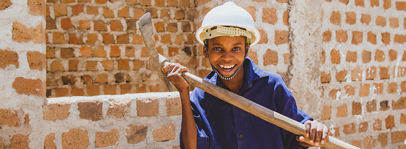 A smiling Tanzanian boy wearing a white construction helmet and holding a pick axe.
