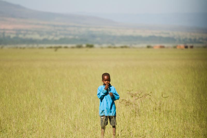 A young boy stands in a grassy field with mountains behind him