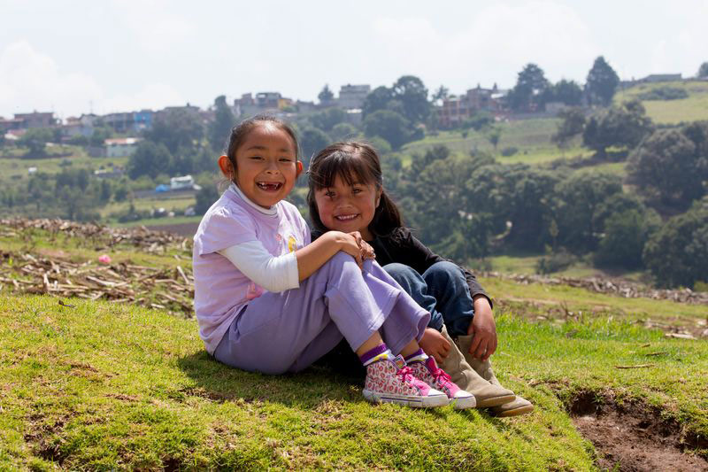 Two girls sit on a hill with mountains behind them