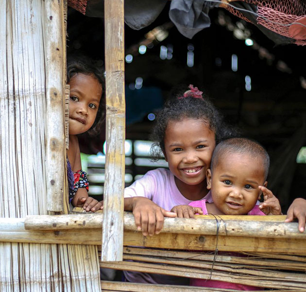 Three children smile while looking out a window in their home.