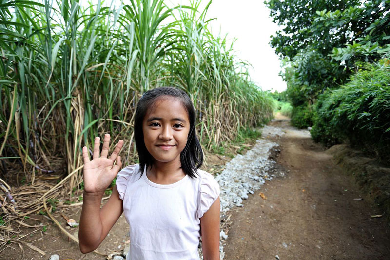 A girl smiles and waves while standing on a dirt path