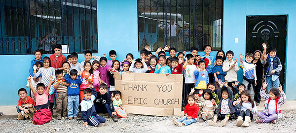 A group of children in front of a blue building hold a cardboard sign with a thank you message written on it
