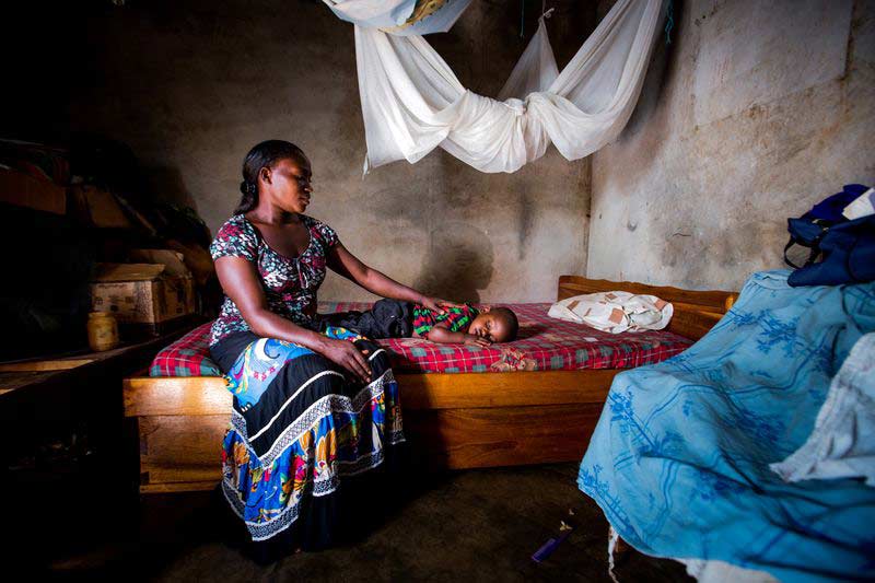 A mother sits with her child on their bed under a mosquito net