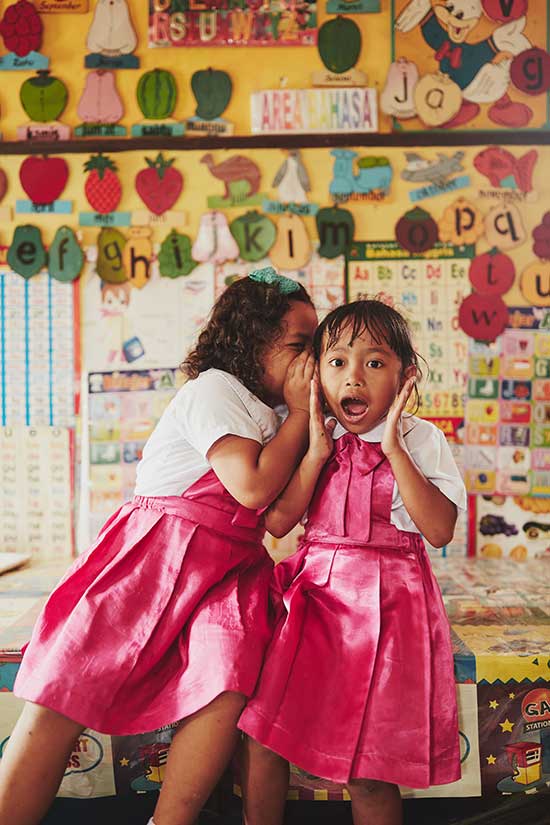A girl in a pink dress whispers in the ear of another girl wearing a pink dress