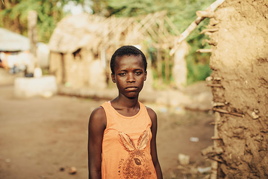 A solemn looking girl with an orange tank top on stands next to a mud hut