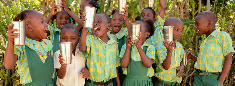 A group of young smiling and laughing Haitian children run hold cups of clean water in the air.