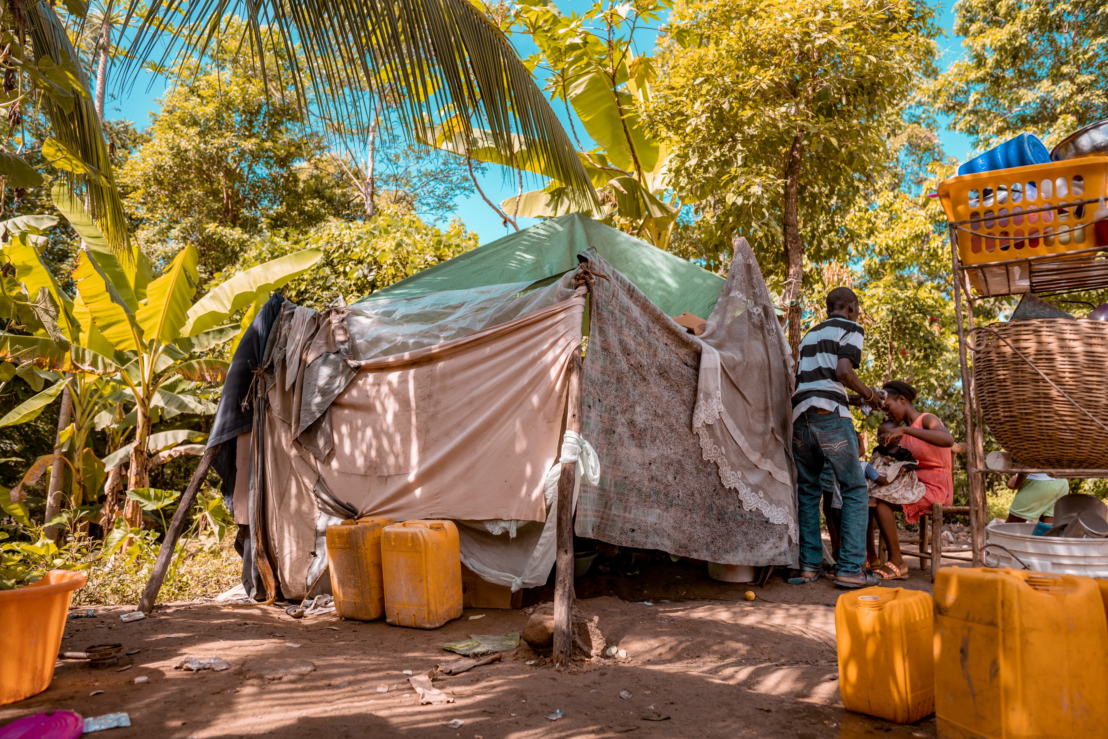 Two adults outdoors surrounded by yellow containers, with a woman holding a baby and a man in a white and blue striped shirt, tropical trees visible in the background.