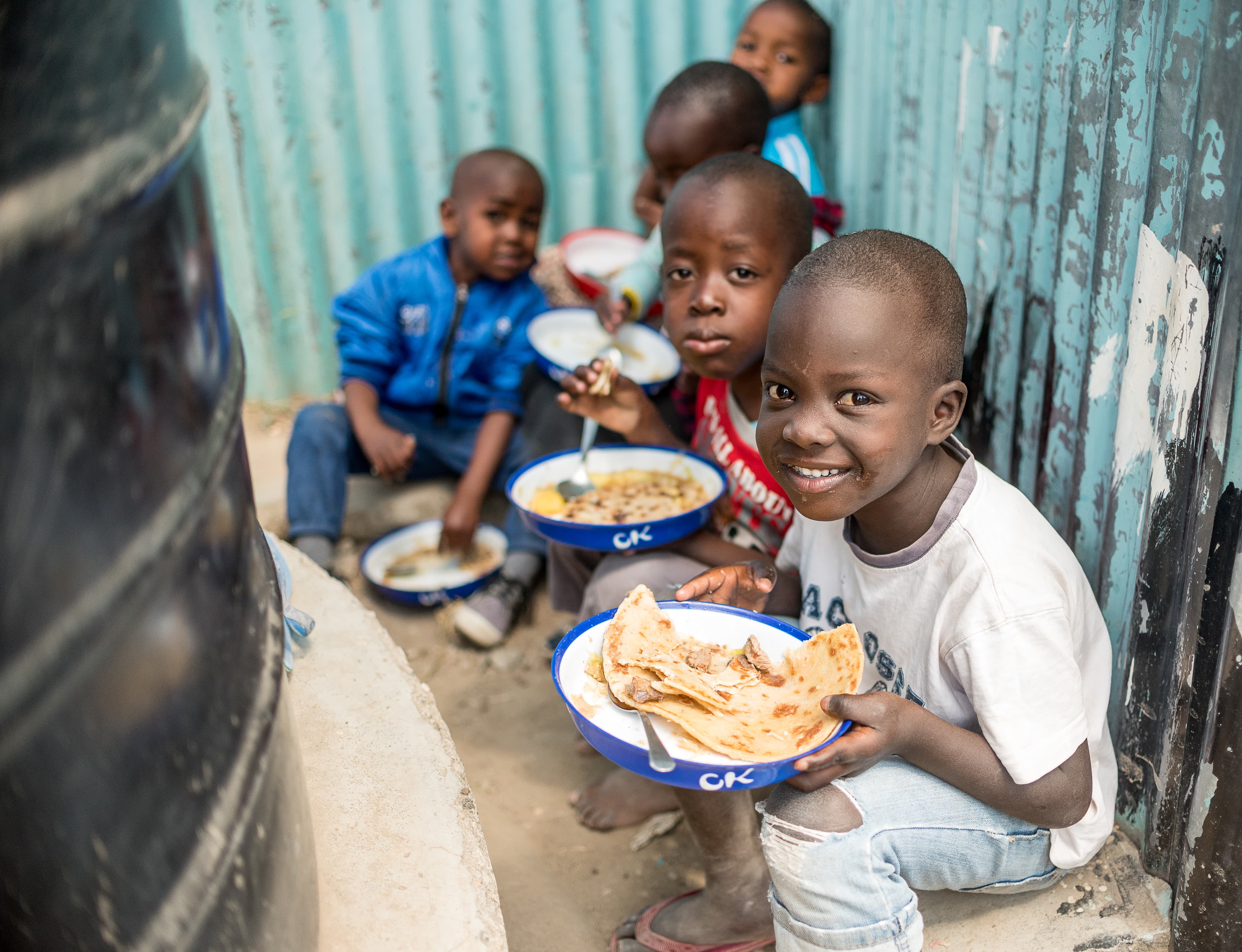 Children in Kenya sit on the ground while holding plates of food.