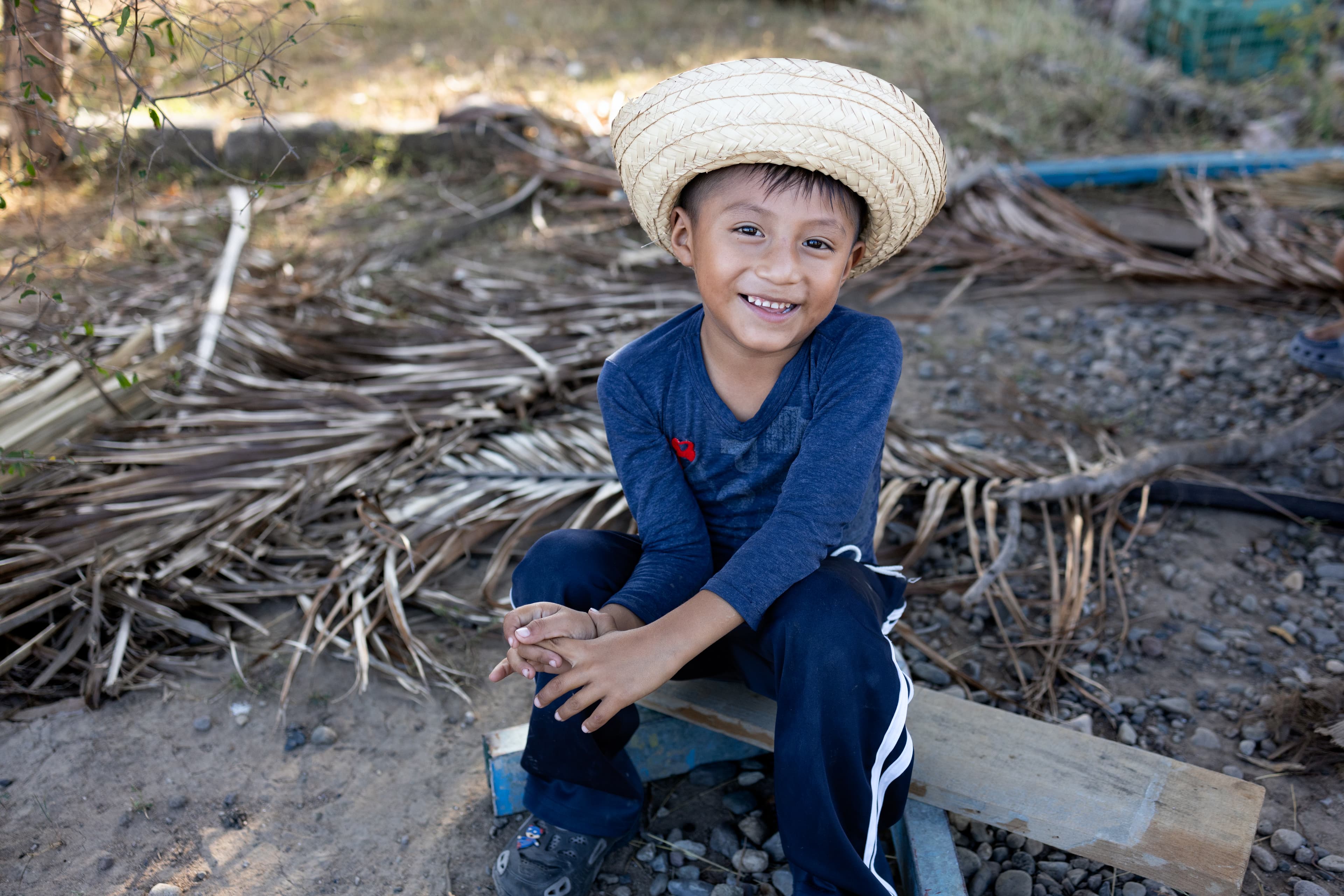 A young Mexican boy sits on a piece of wood while wearing a straw hat and smiling.