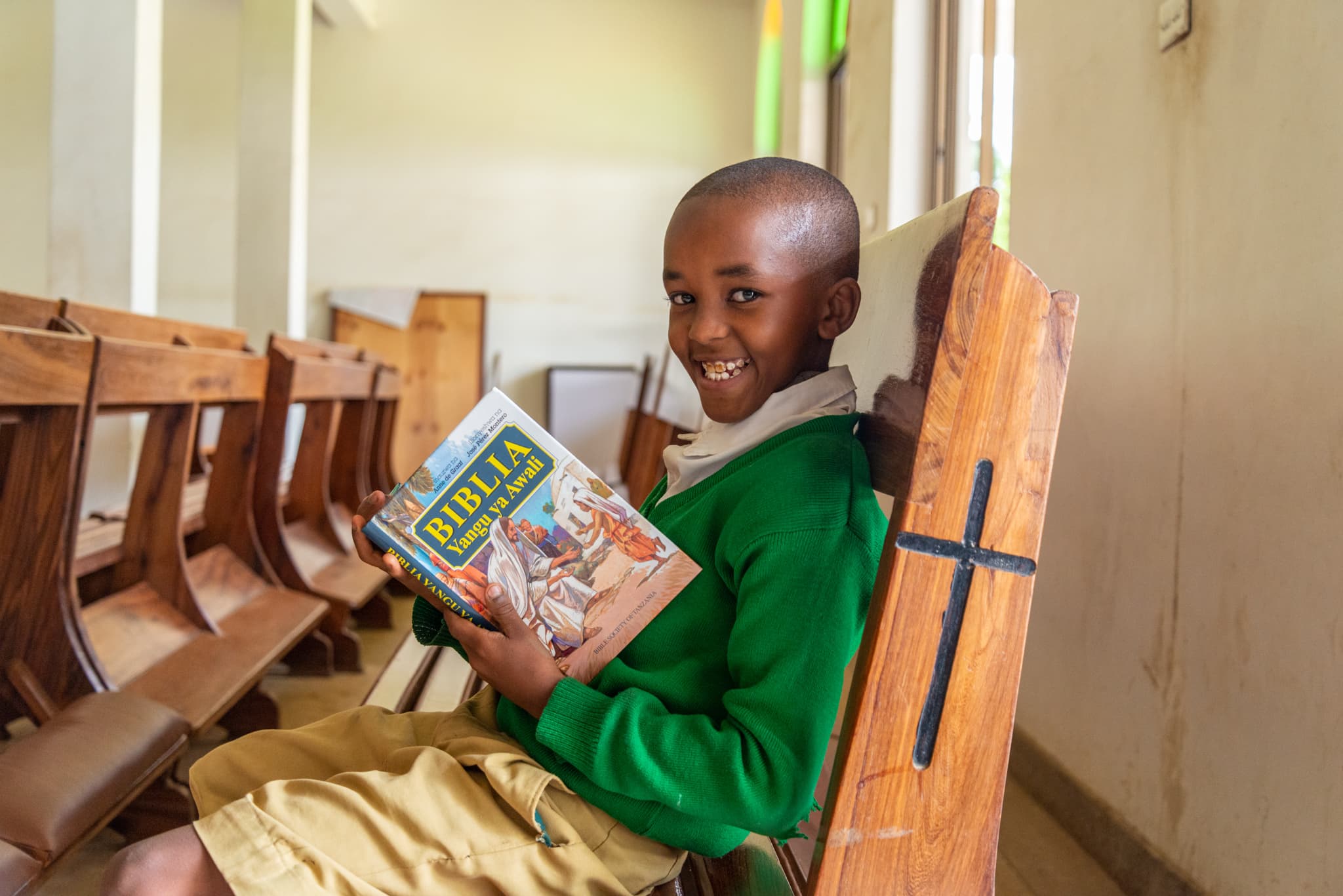 An African child sits in a wooden pew and smiles while holding an open children’s Bible.