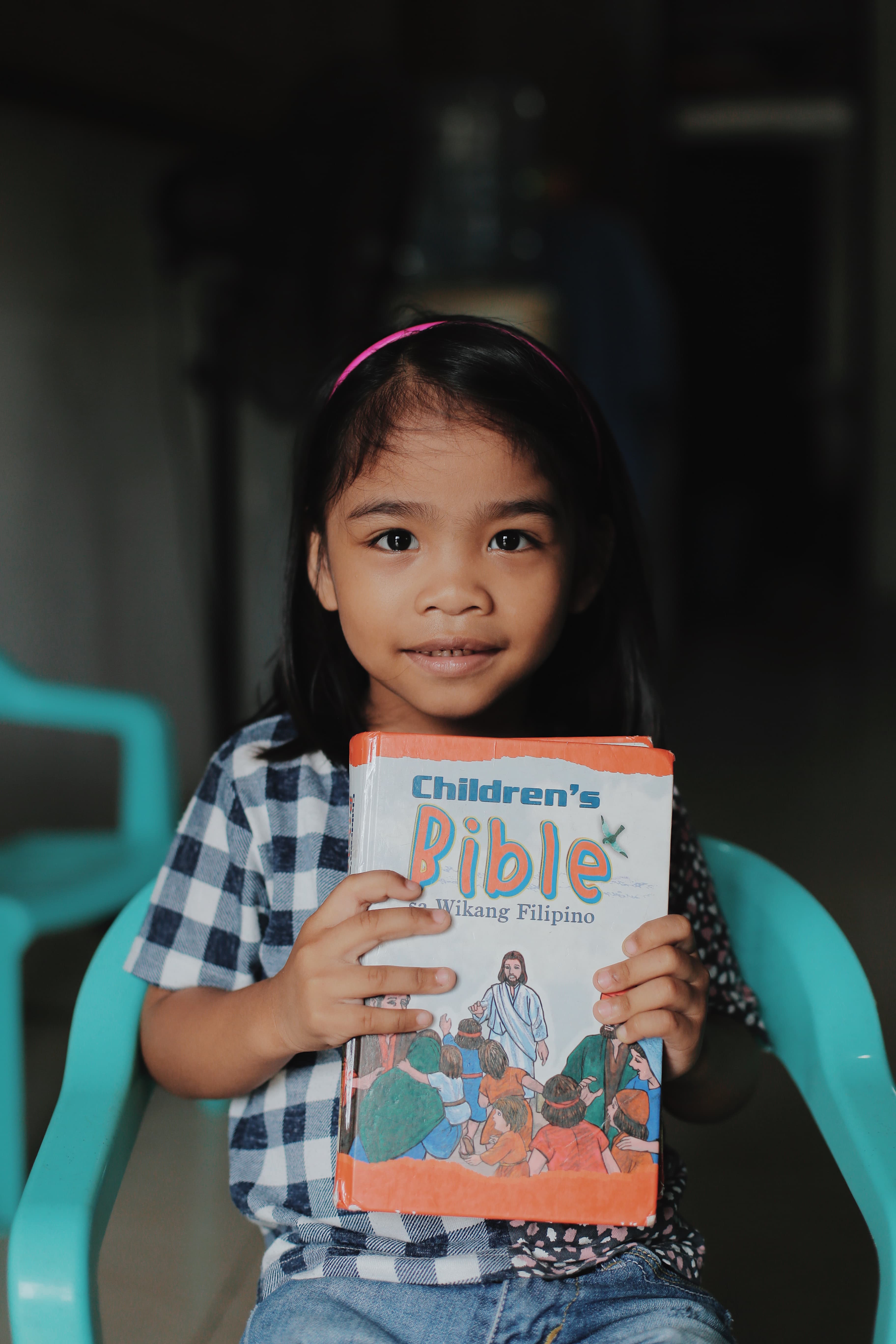 A little girl wearing a checkered shirt sits in a chair holding a children's Bible and smiles