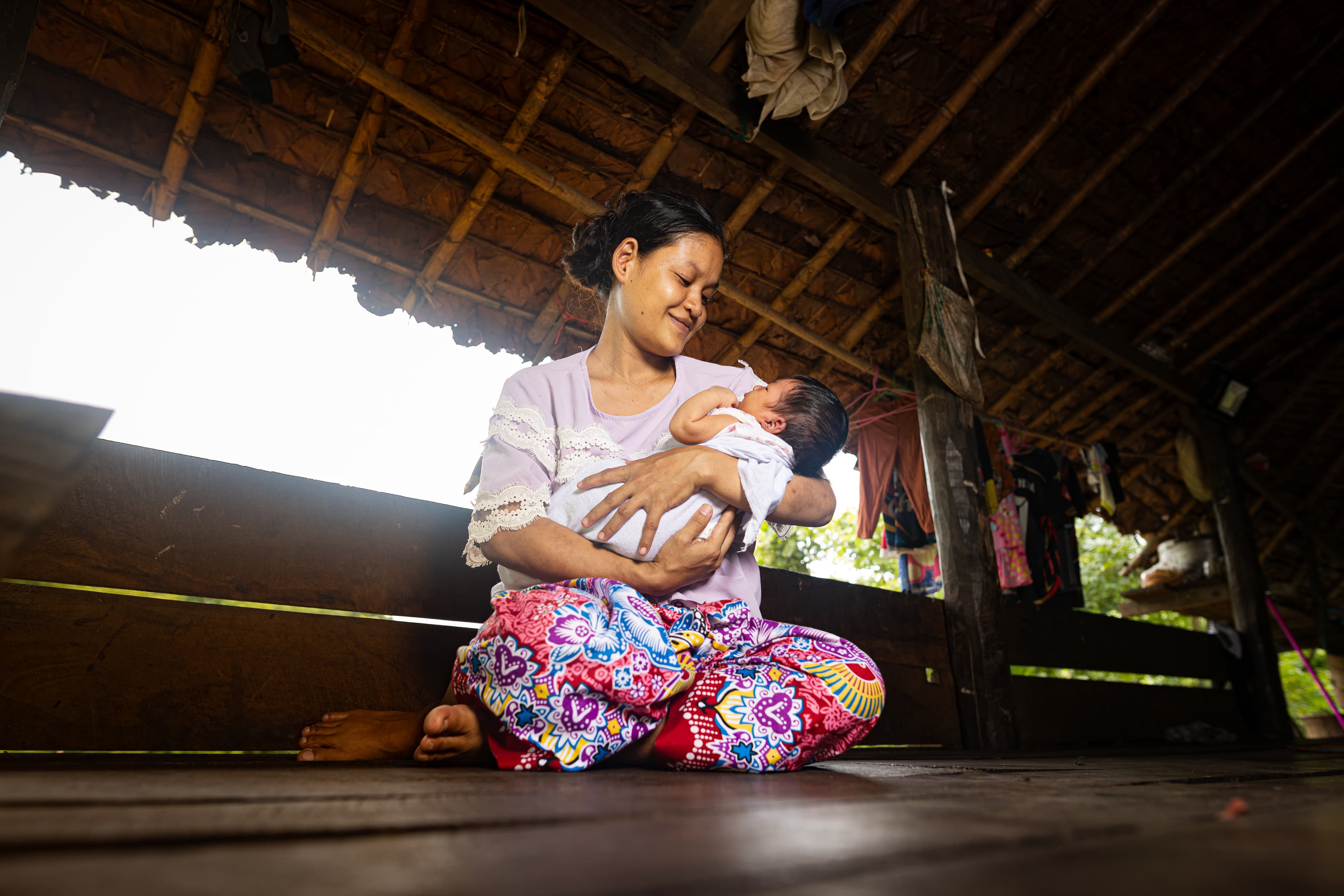 Thai woman sits on the floor inside her home, smiling while holding her baby.