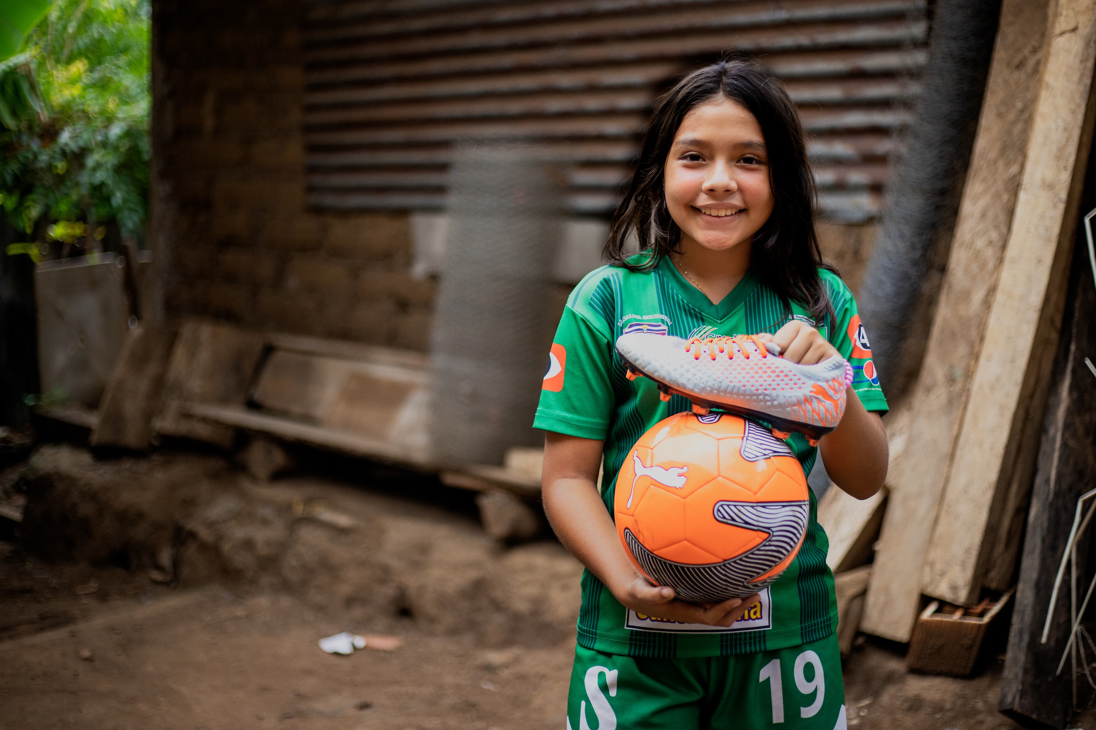 A young girl stands holding a soccer cleat and a soccer ball while smiling.