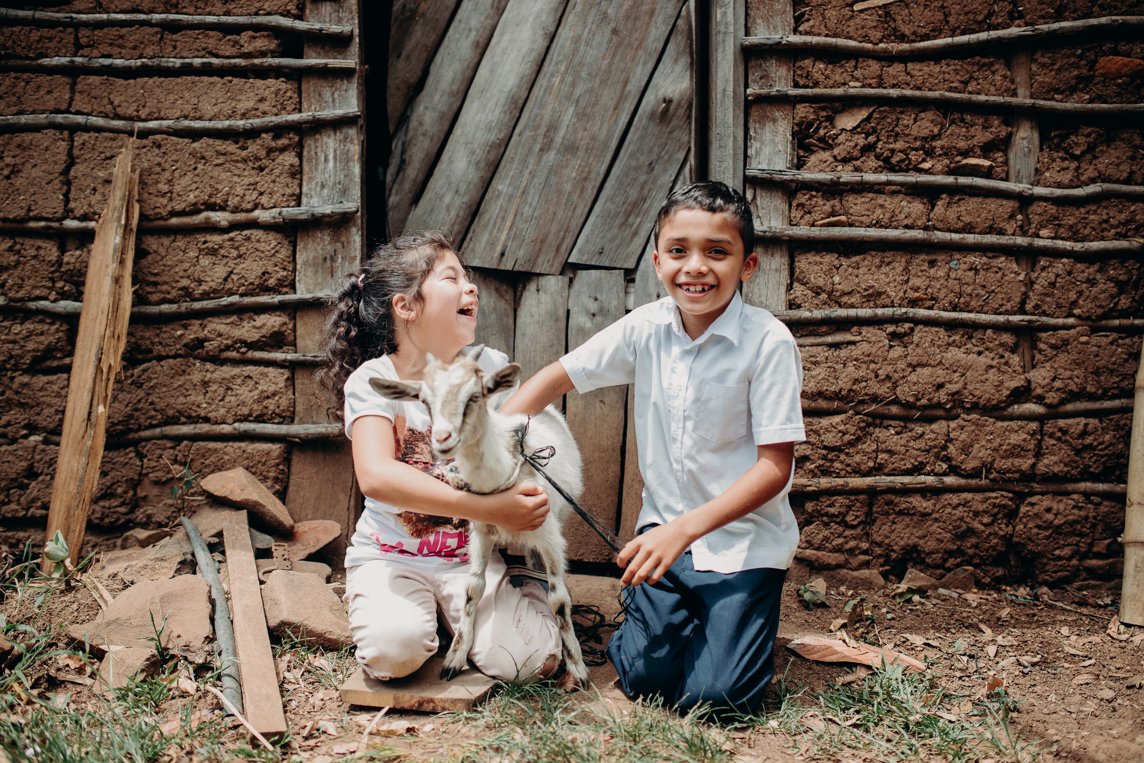 A young boy and girl kneel on the ground in front of a wood and mud building, holding a goat and laughing.