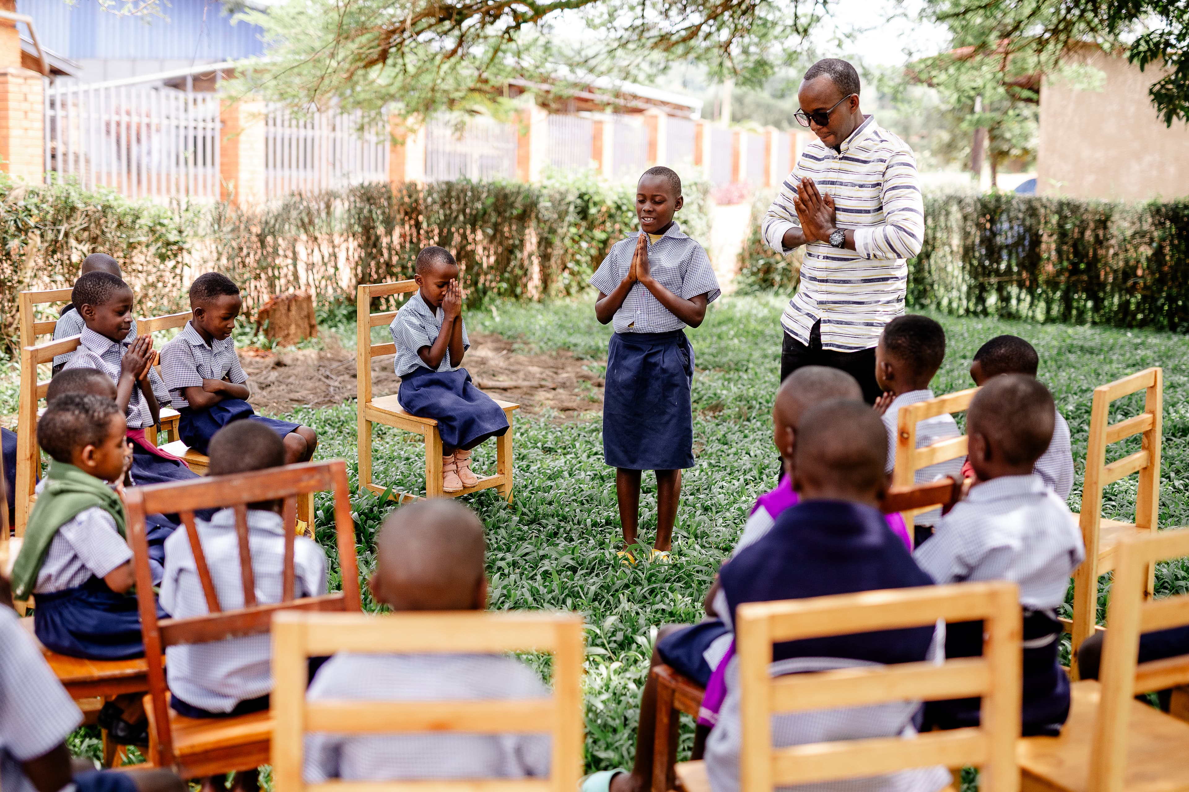 An African man stands in a circle of children sitting in chairs.