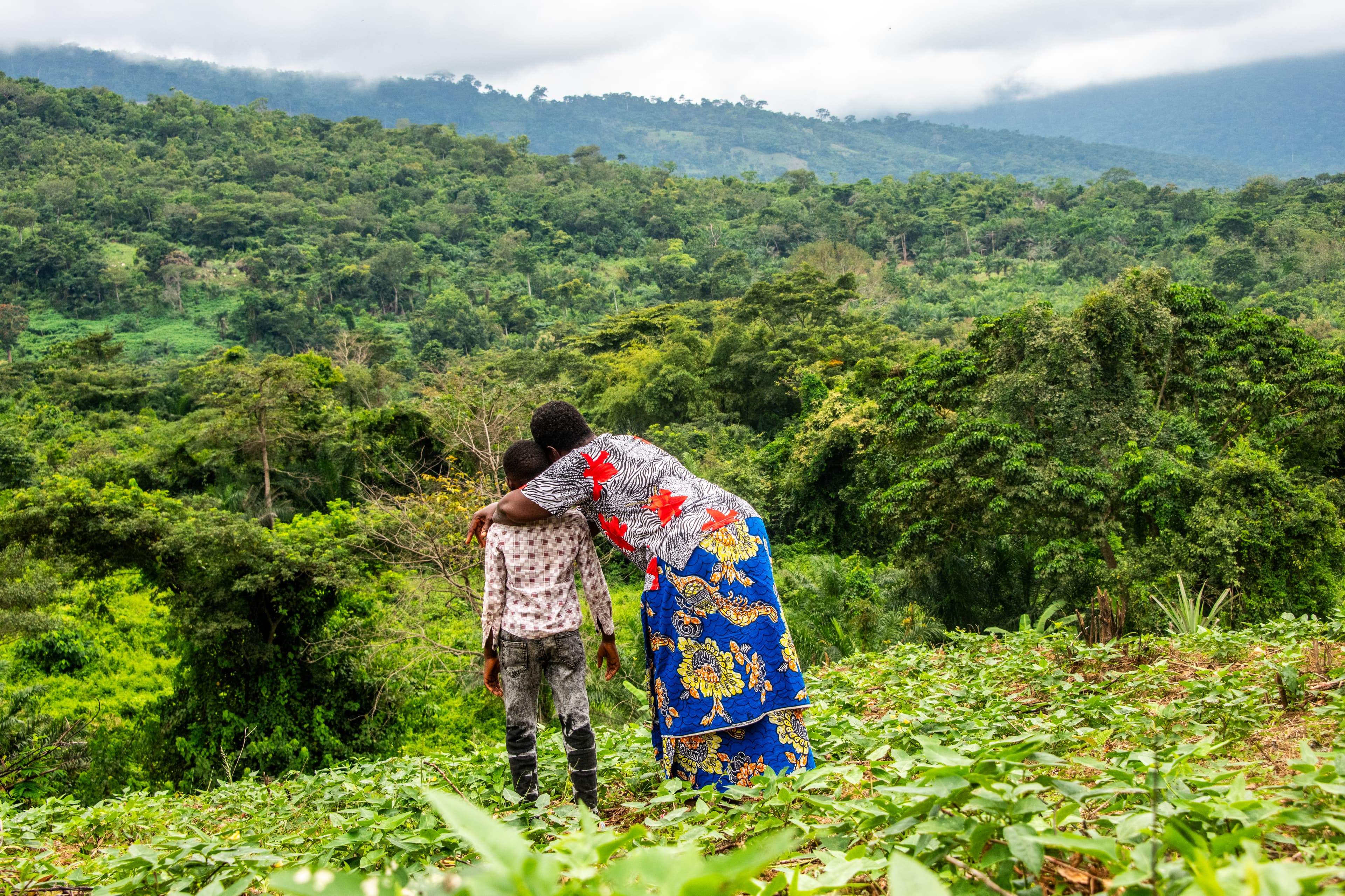 A mother and son are standing on top of a hill in their neighborhood as the mother bends down to hug her son.
