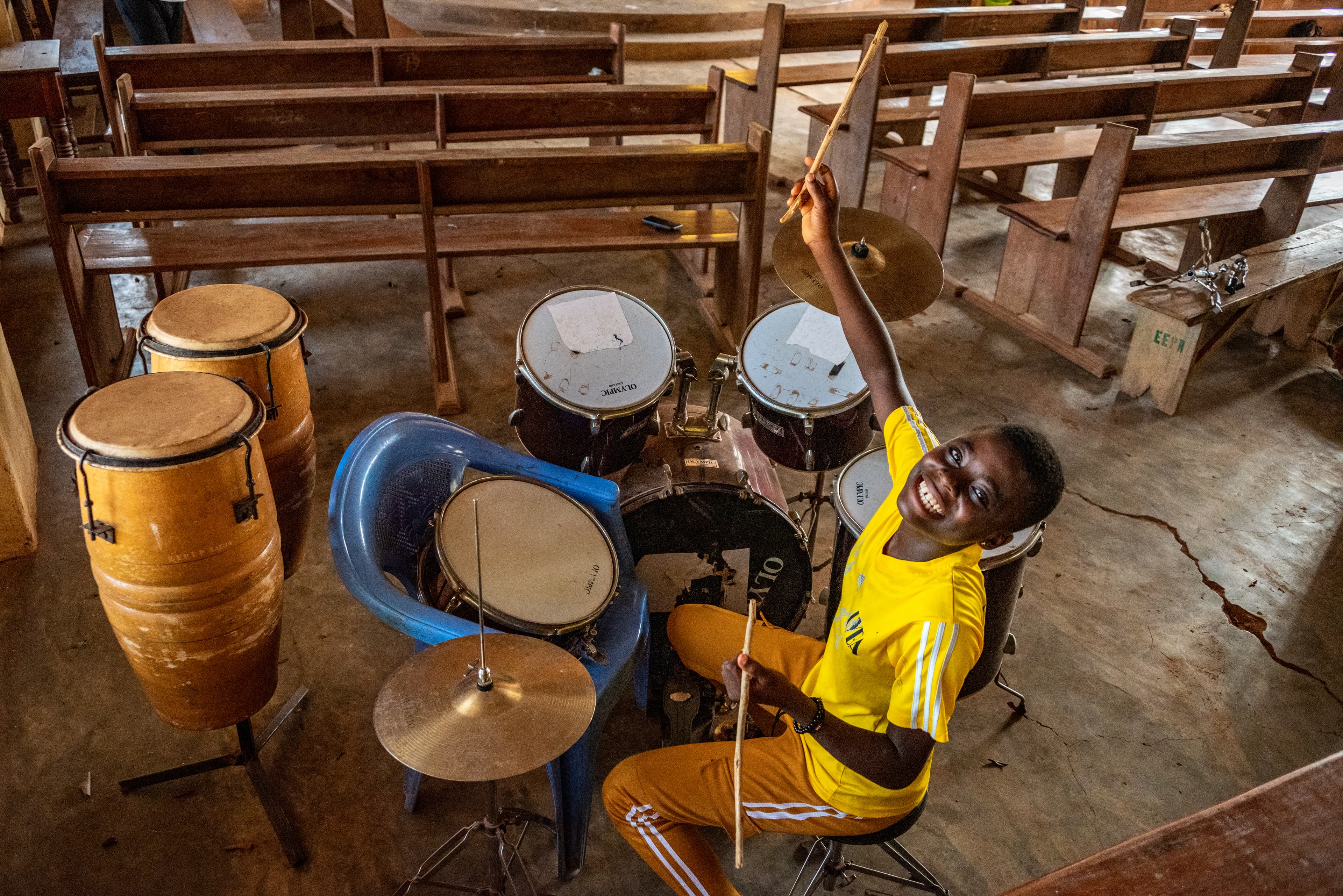 A young boy sits at a set of drums and smiles while holding drumsticks in the air.
