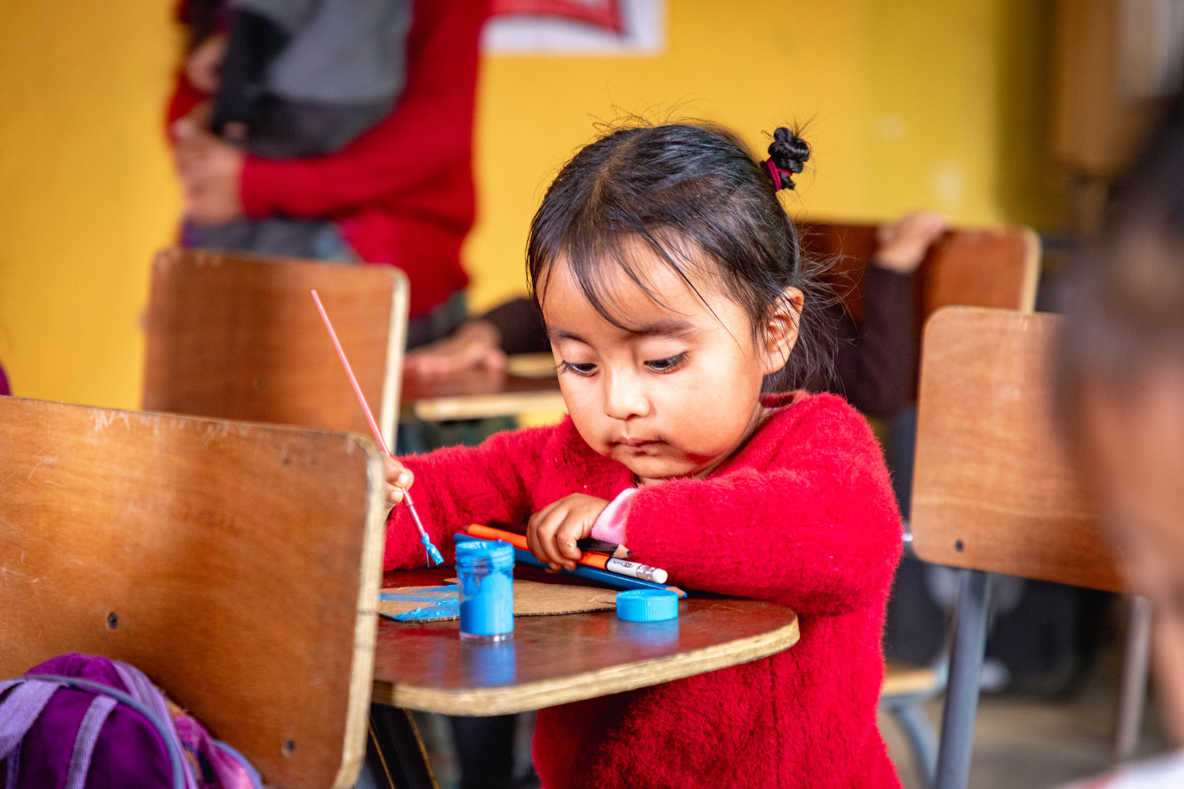A young girl sits at a wooden school desk while painting.