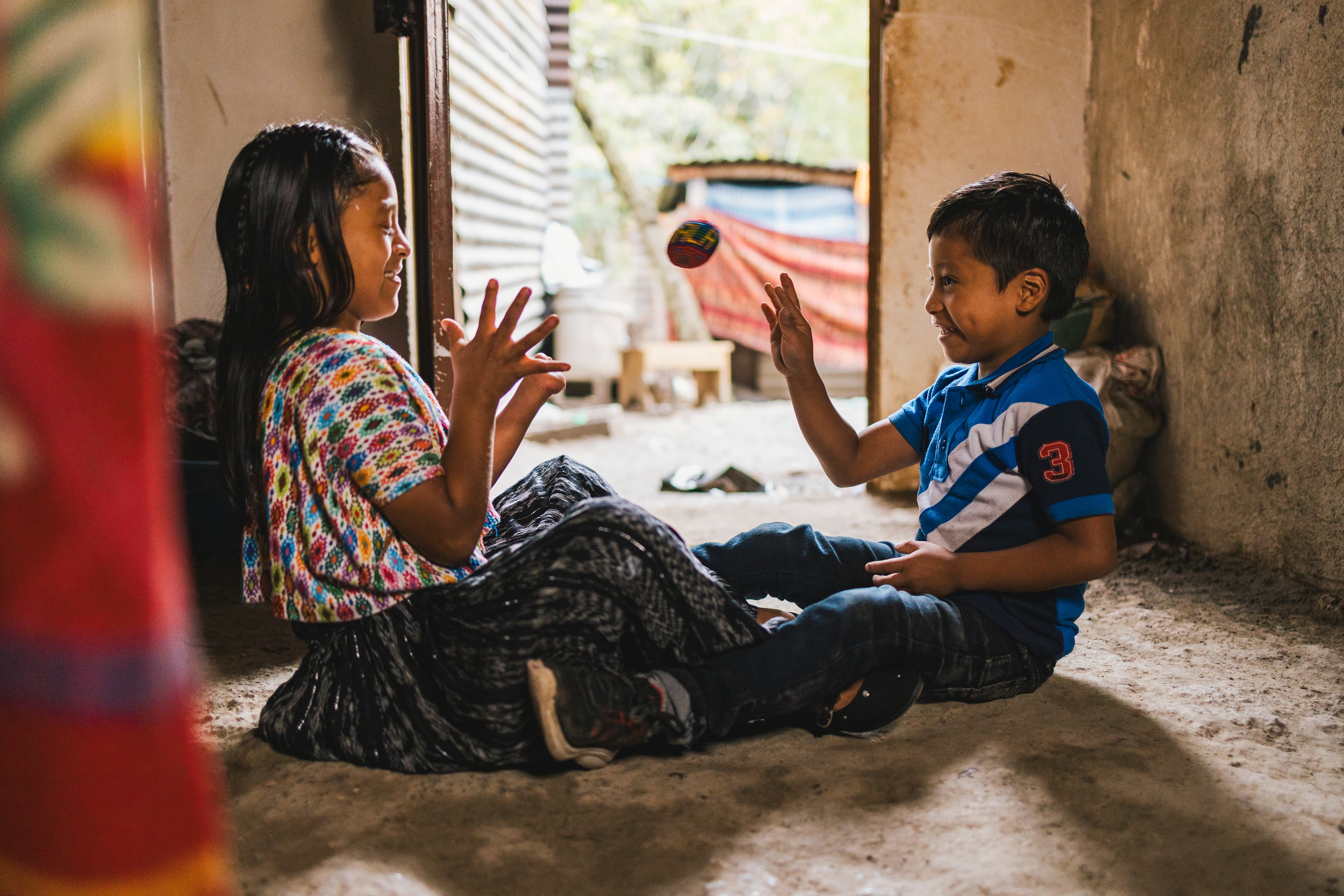 A girl and younger boy toss a knit ball back and forth while sitting on a dirt floor. The girl is wearing a colorful patterned shirt and the boy is in a blue and white polo shirt.