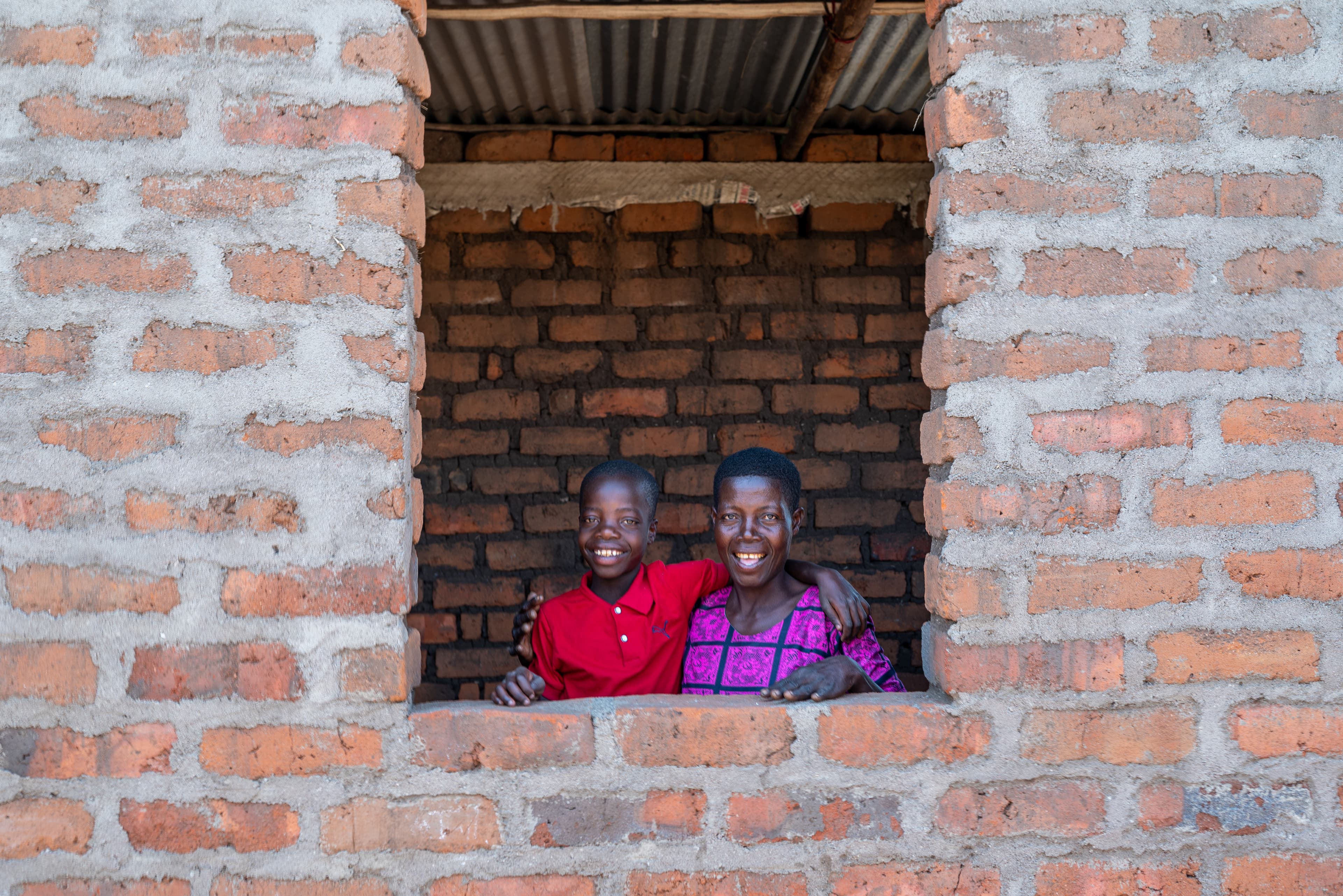 A boy wearing a red shirt stands in the window of his new home with his arm around his mother, wearing a purple dress.