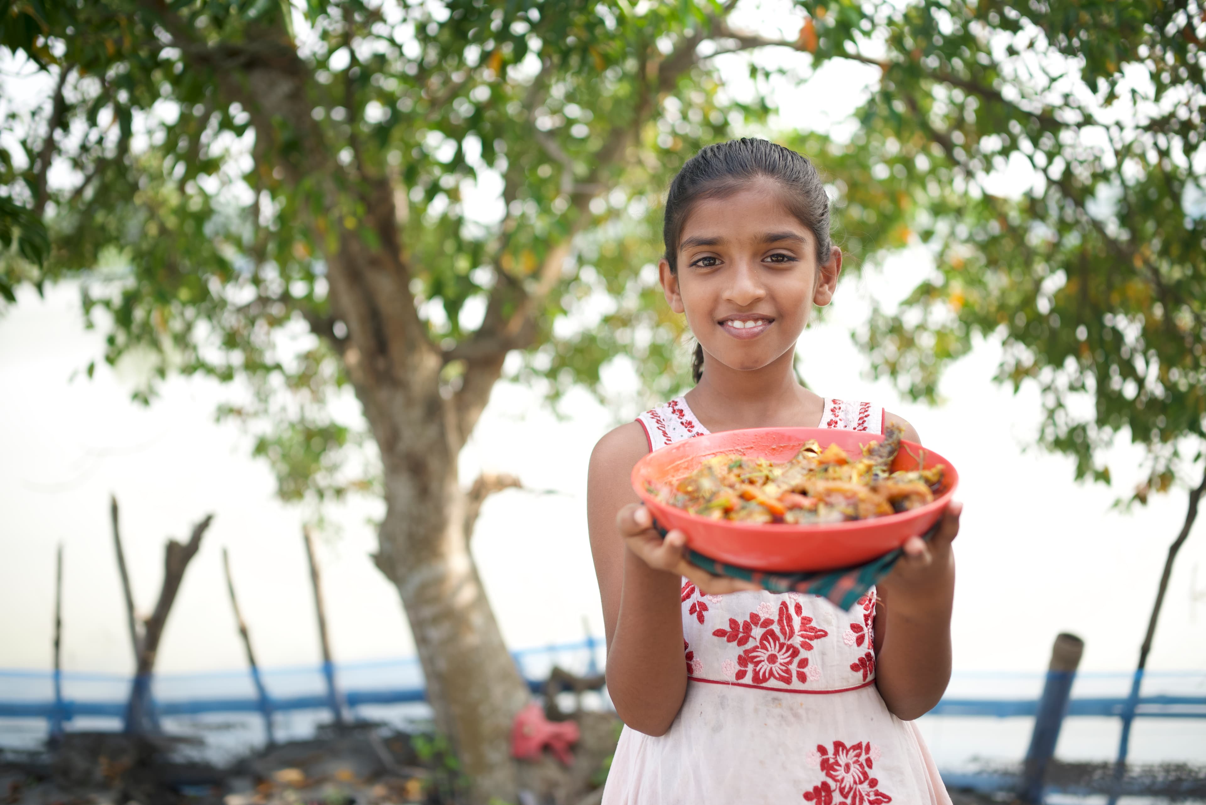 A young girl smiles for the camera while holding a red bowl of her favorite meal.