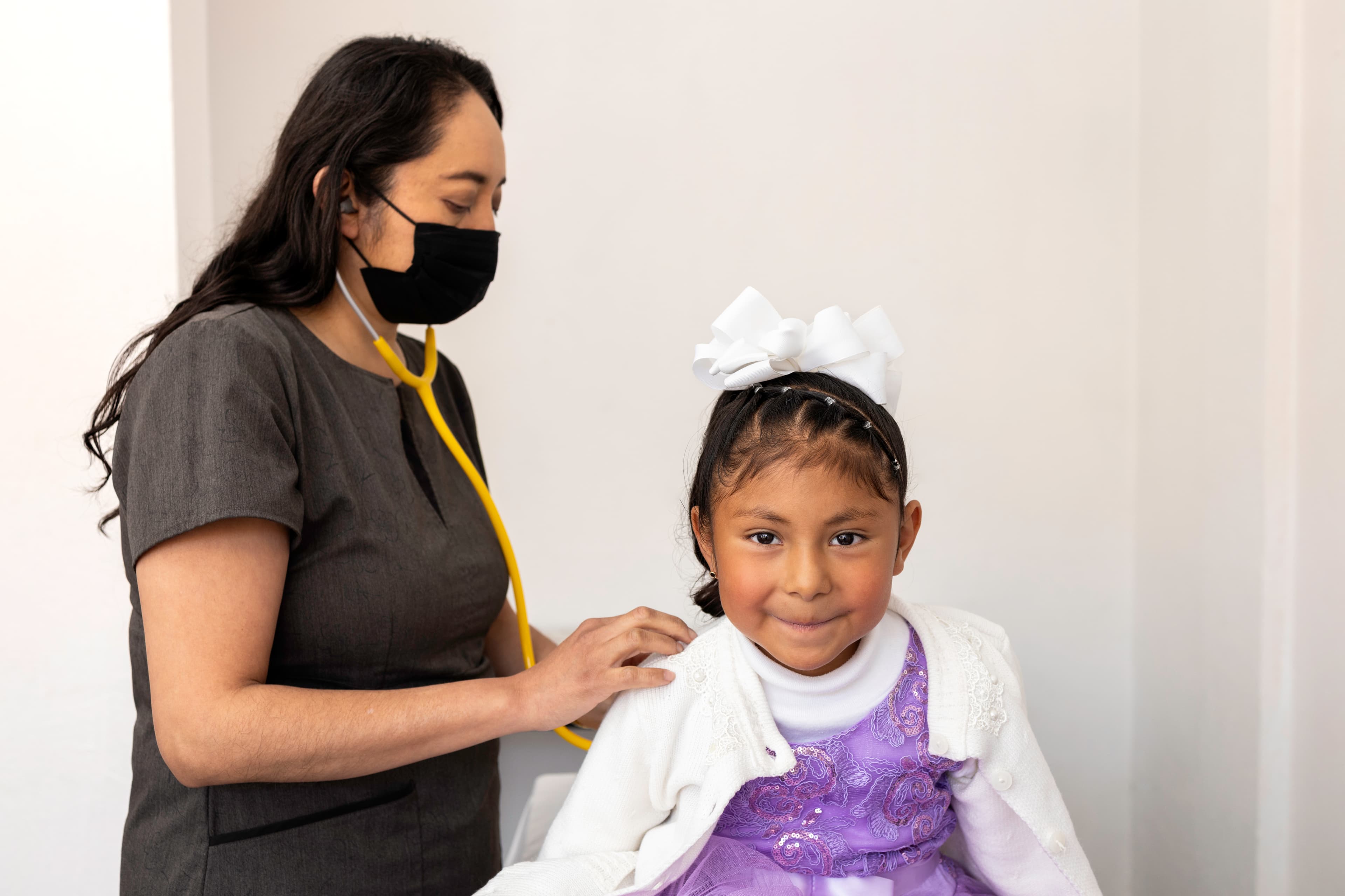 A young girl in a purple dress gets her heart checked by a nurse with a stethoscope.