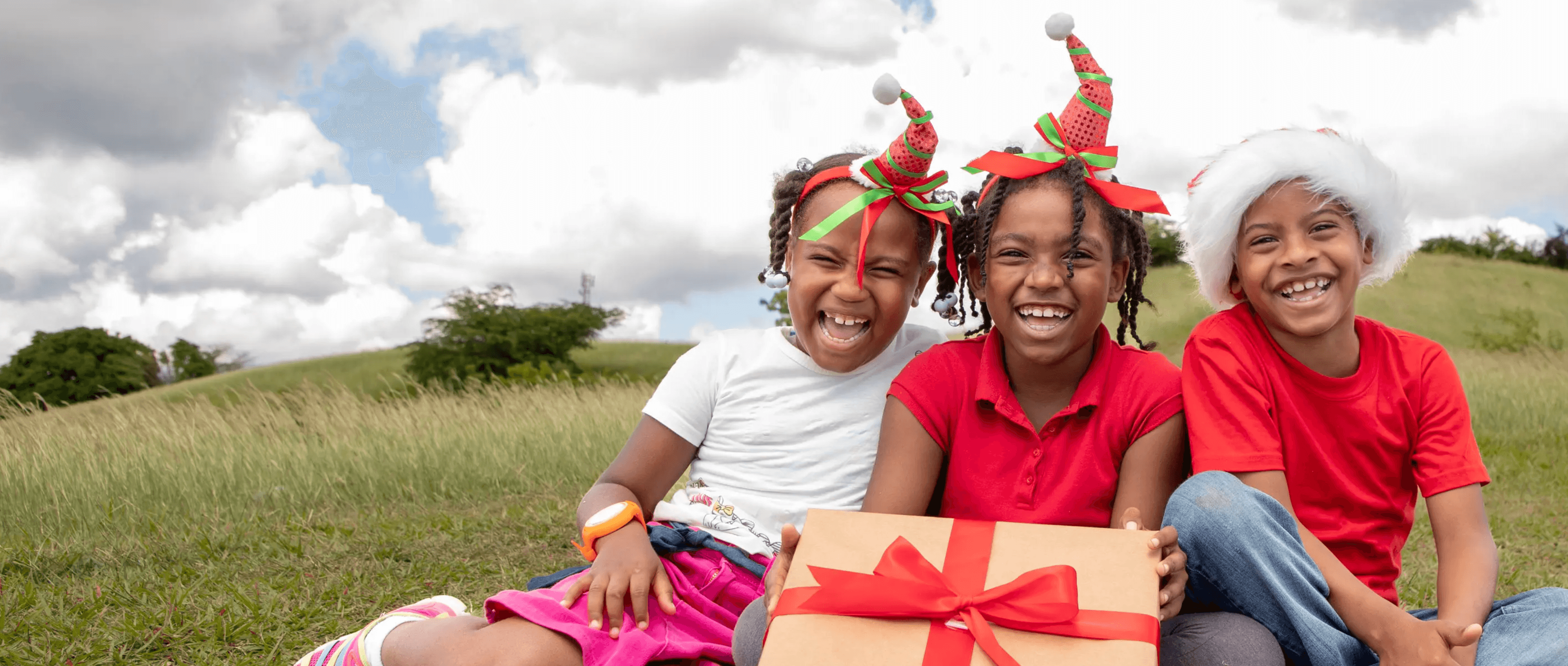 Three children are sitting on a grassy hill holding a large Christmas gift.