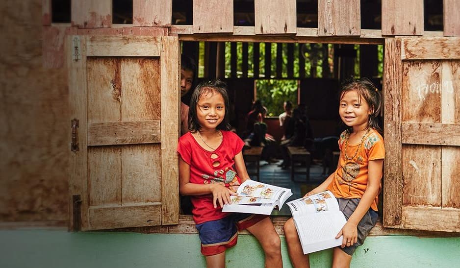 Two playful African boys embrace outside their school; one wearing a yellow shirt, the other wearing a blue shirt.