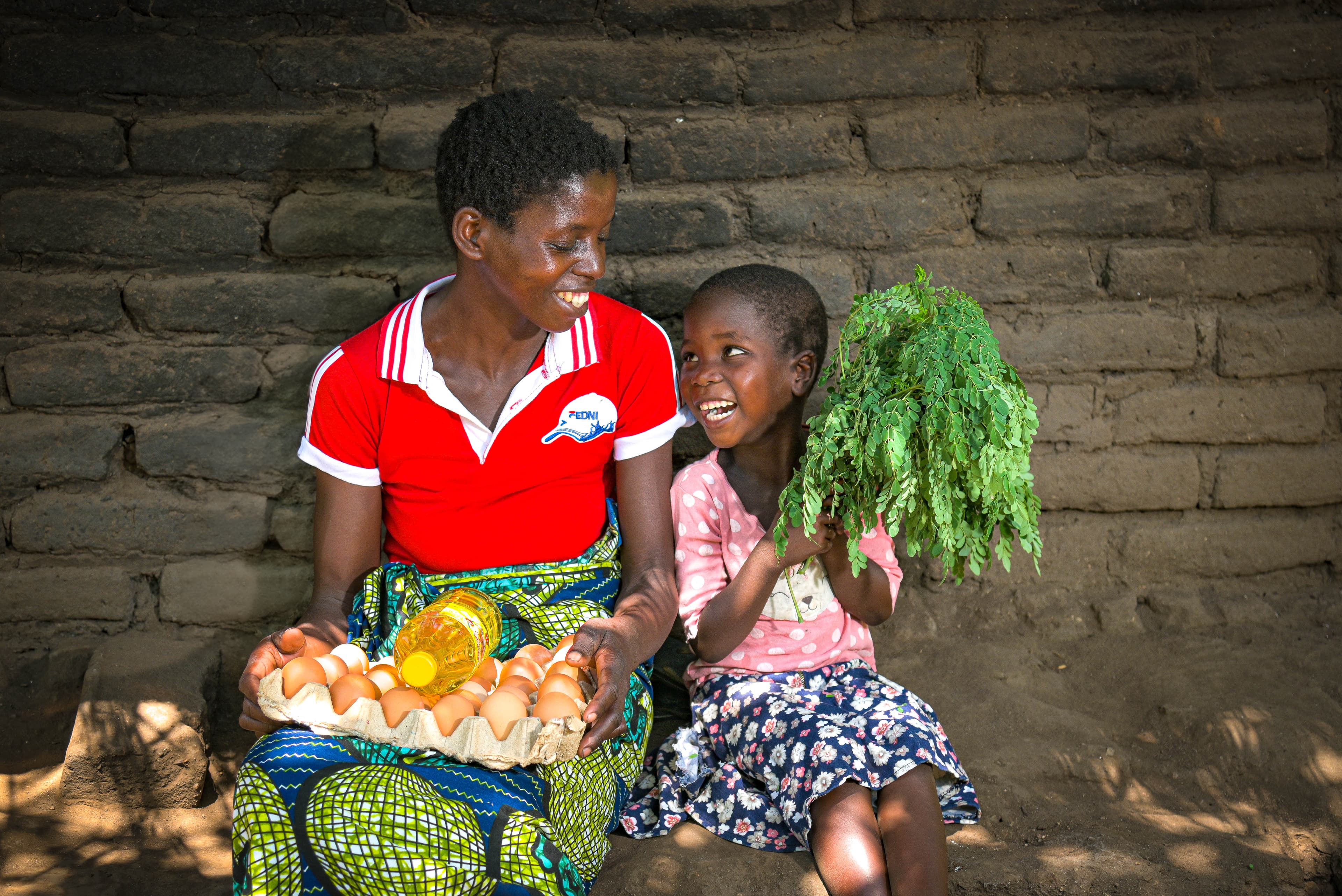 An African woman and a young African girl sit next to eat other holding eggs and moringa leaves.