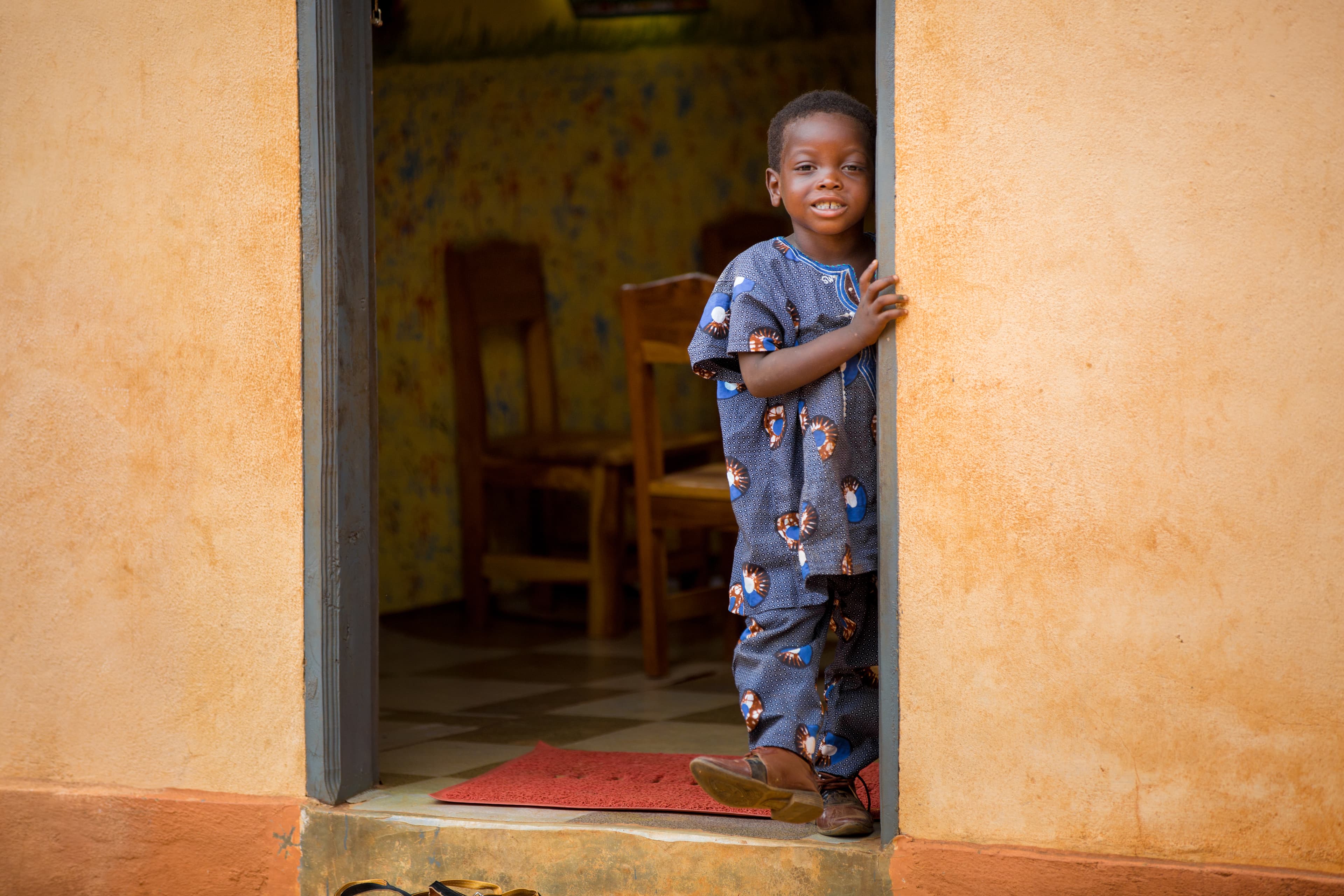 A young boy wearing a patterned blue shirt and pants stands in the doorway of an orange concrete building.