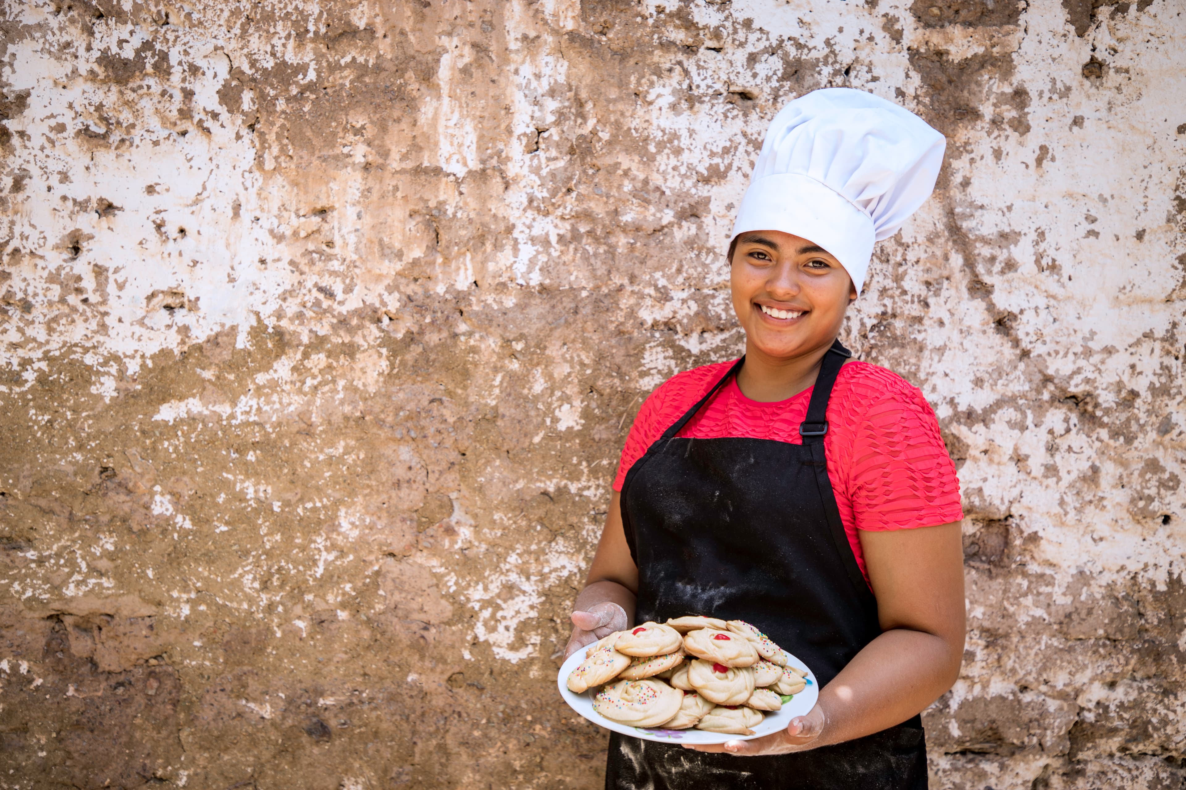 A young women wearing a black apron and white chef’s hat smiles while holding a plate of cookies.