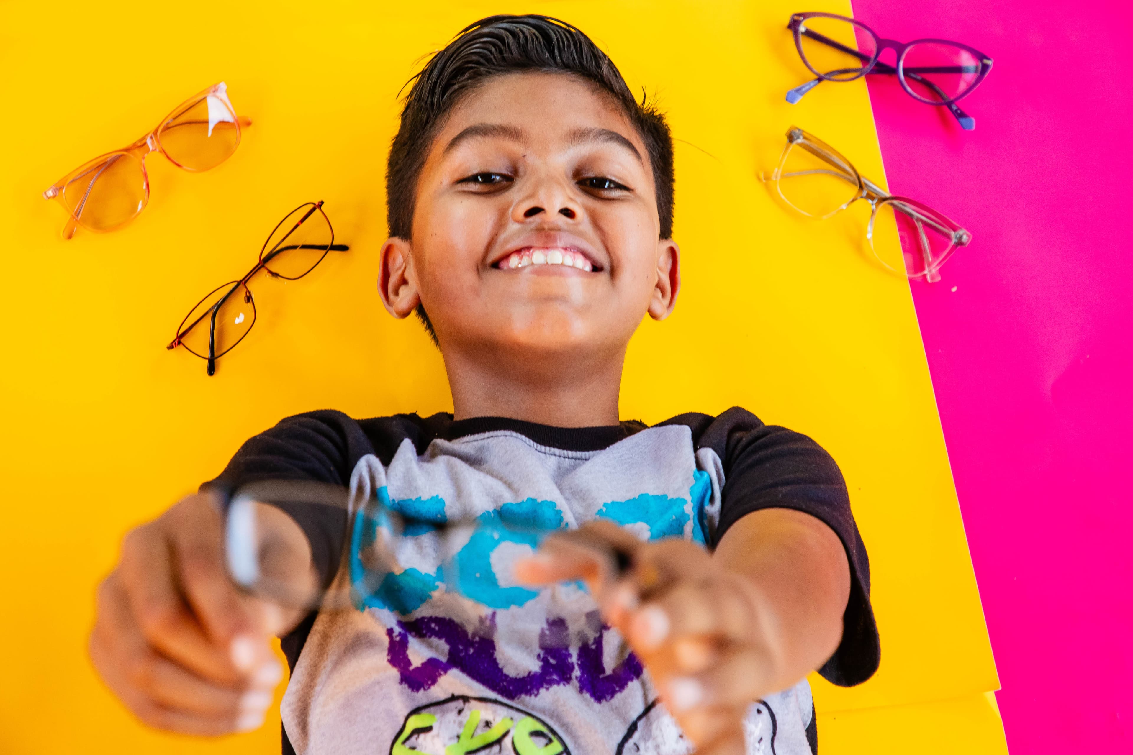 A young boy laying on a bright yellow background holds glasses up the camera and smiles.