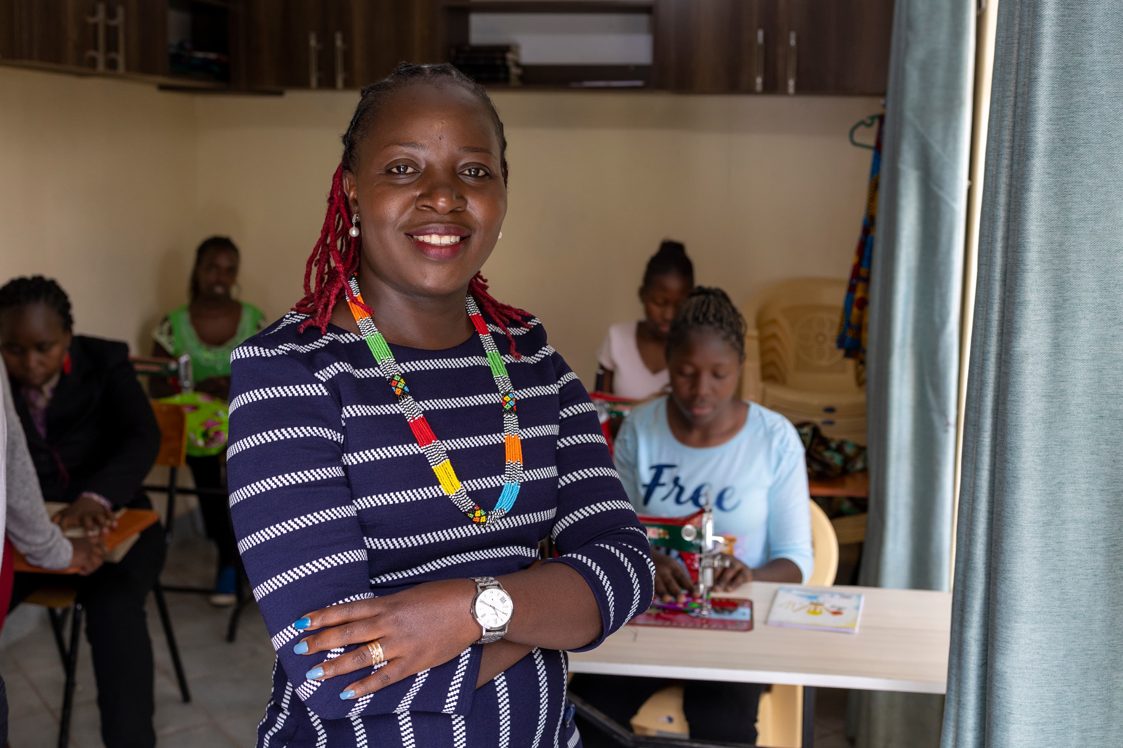 A woman wearing a striped dress and a brightly colored necklace stands with her arms crossed while smiling for the camera.