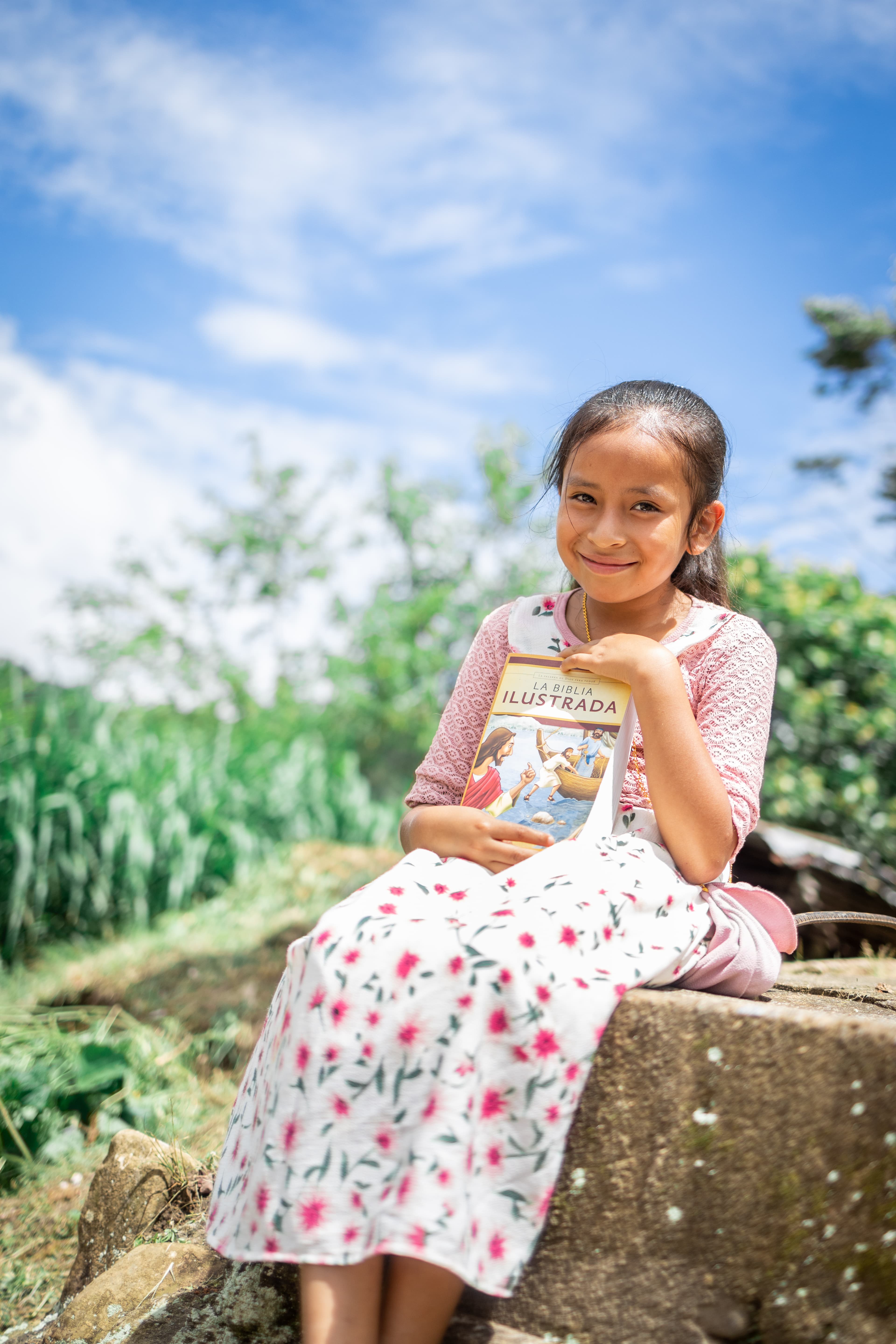 Peruvian girl wearing a floral dress sits on a cement wall while holding a Bible.
