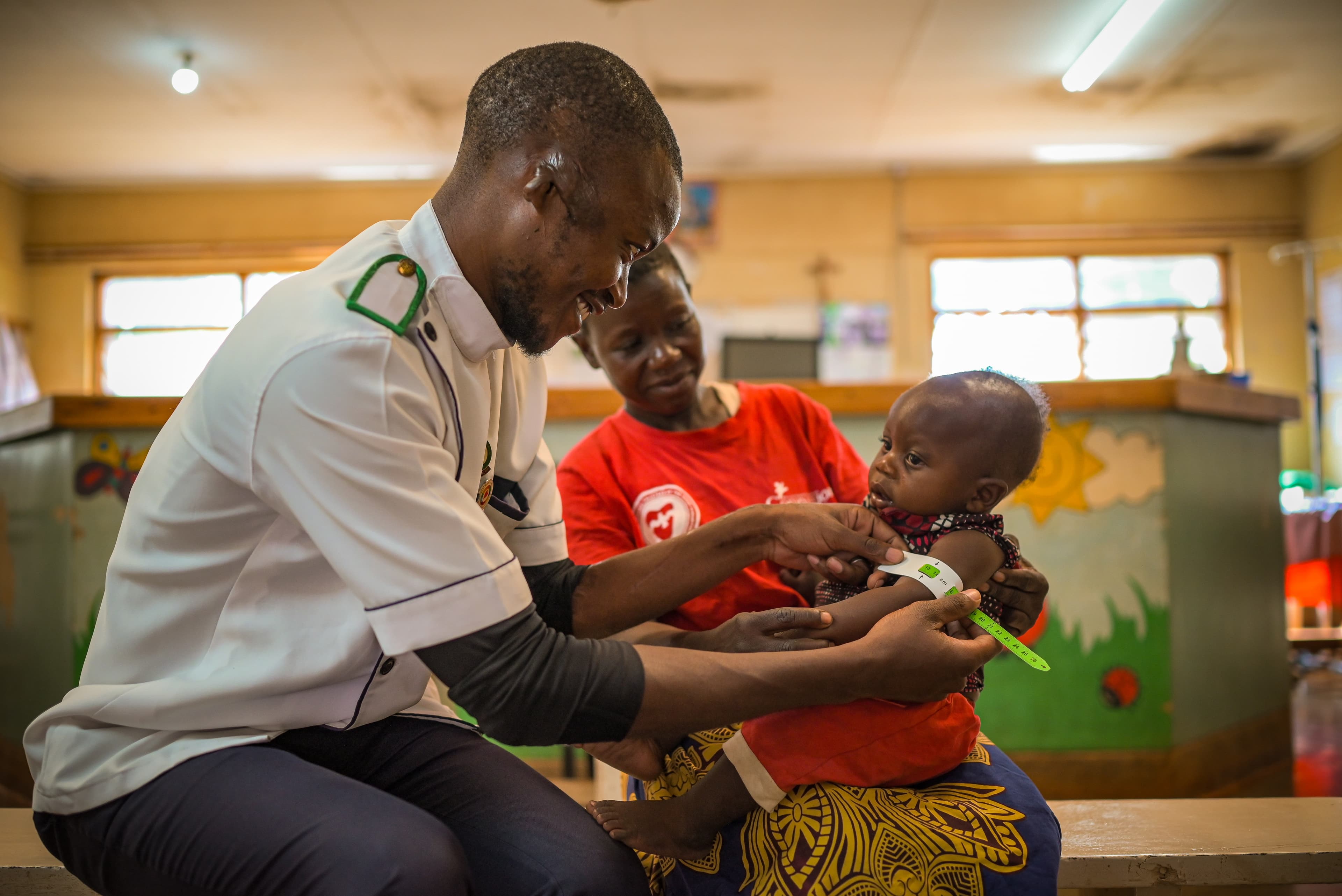 An infant African girl is measured by a smiling doctor as she sits in her mother's lap.