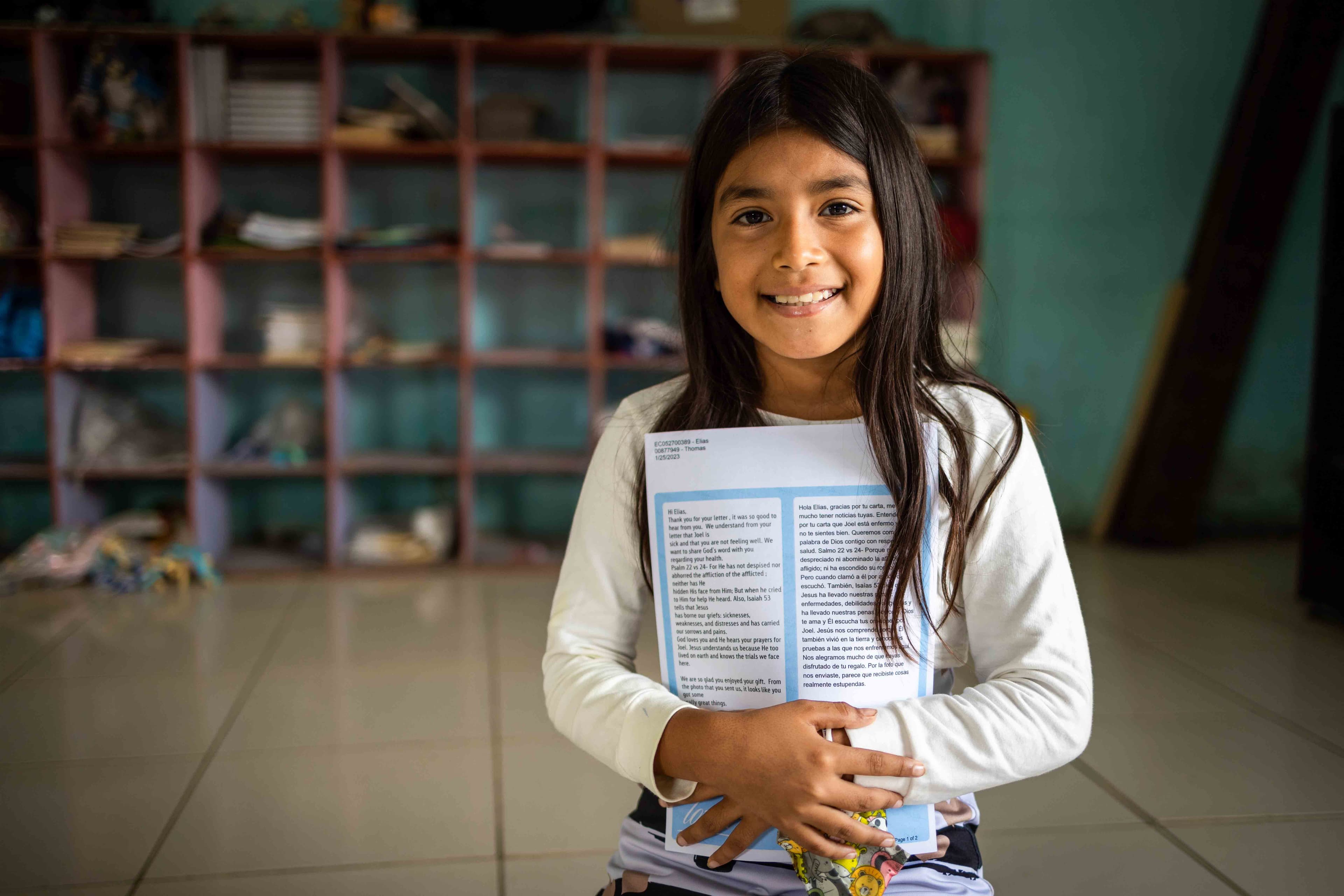A young girl is standing hugging a sponsor letter as she smiles.