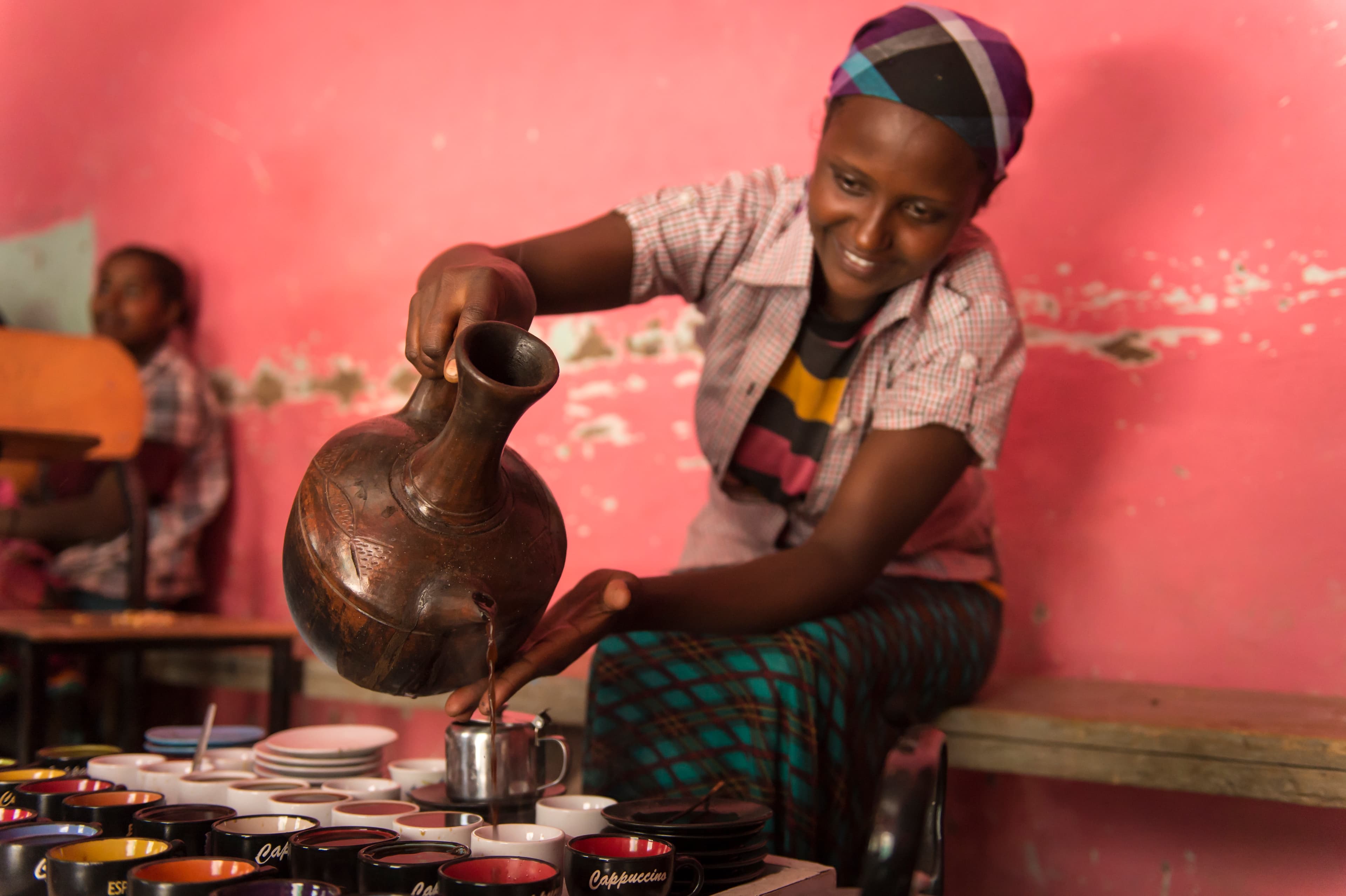 An older woman pours coffee from a jug while smiling.