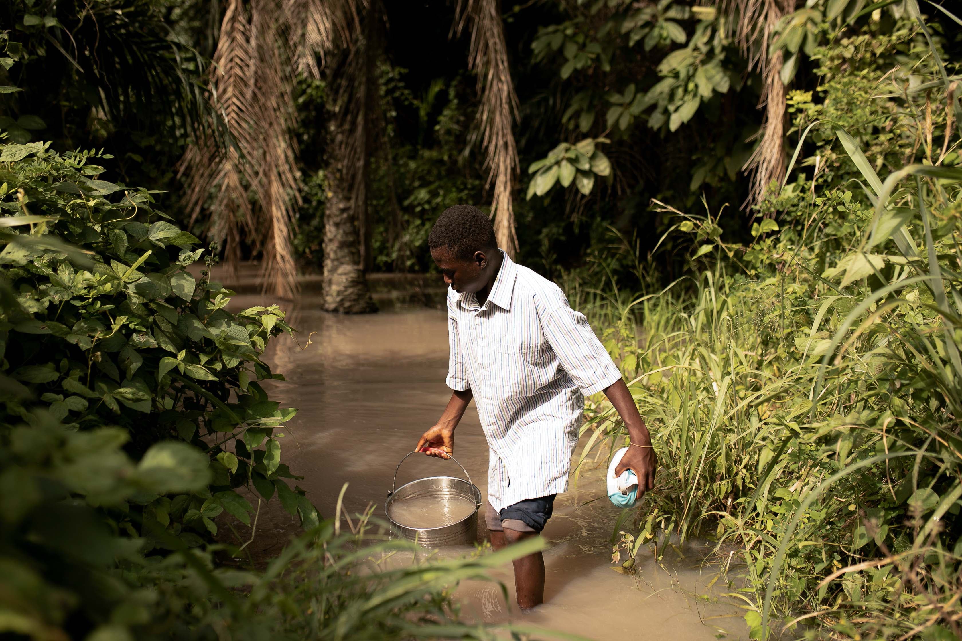 A teen African boy gathers water in a bucket from a muddy stream.