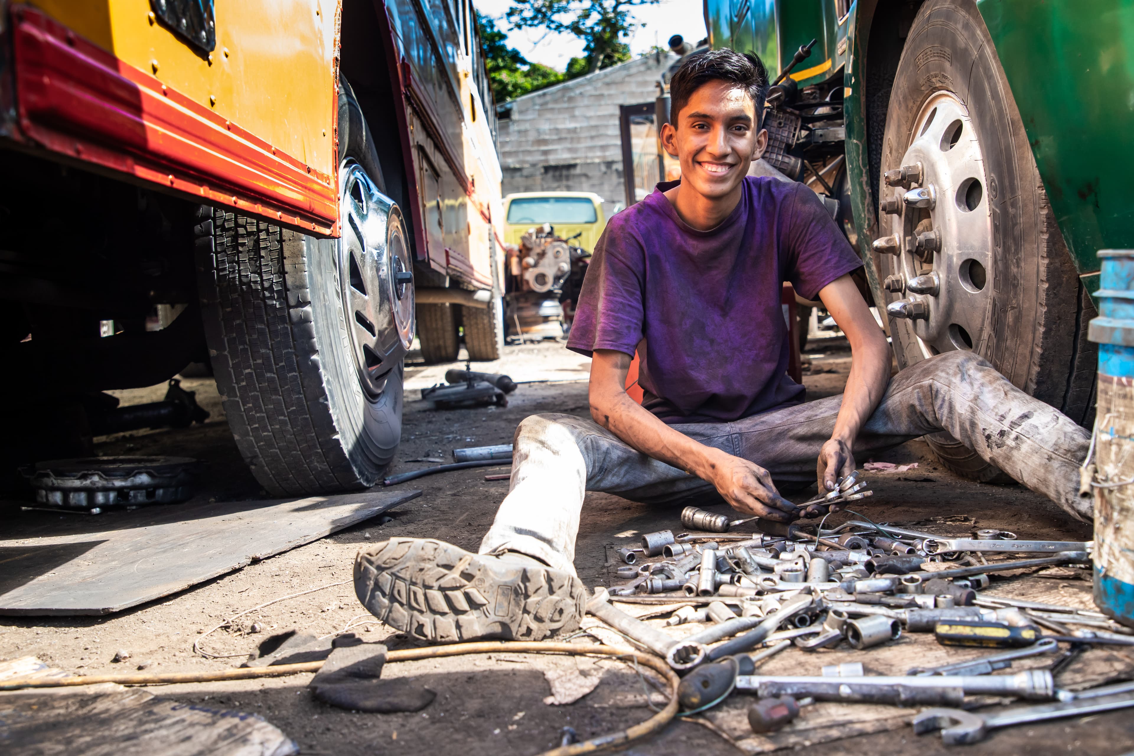 A young man sits between two vehicles surrounded by tools while smiling.