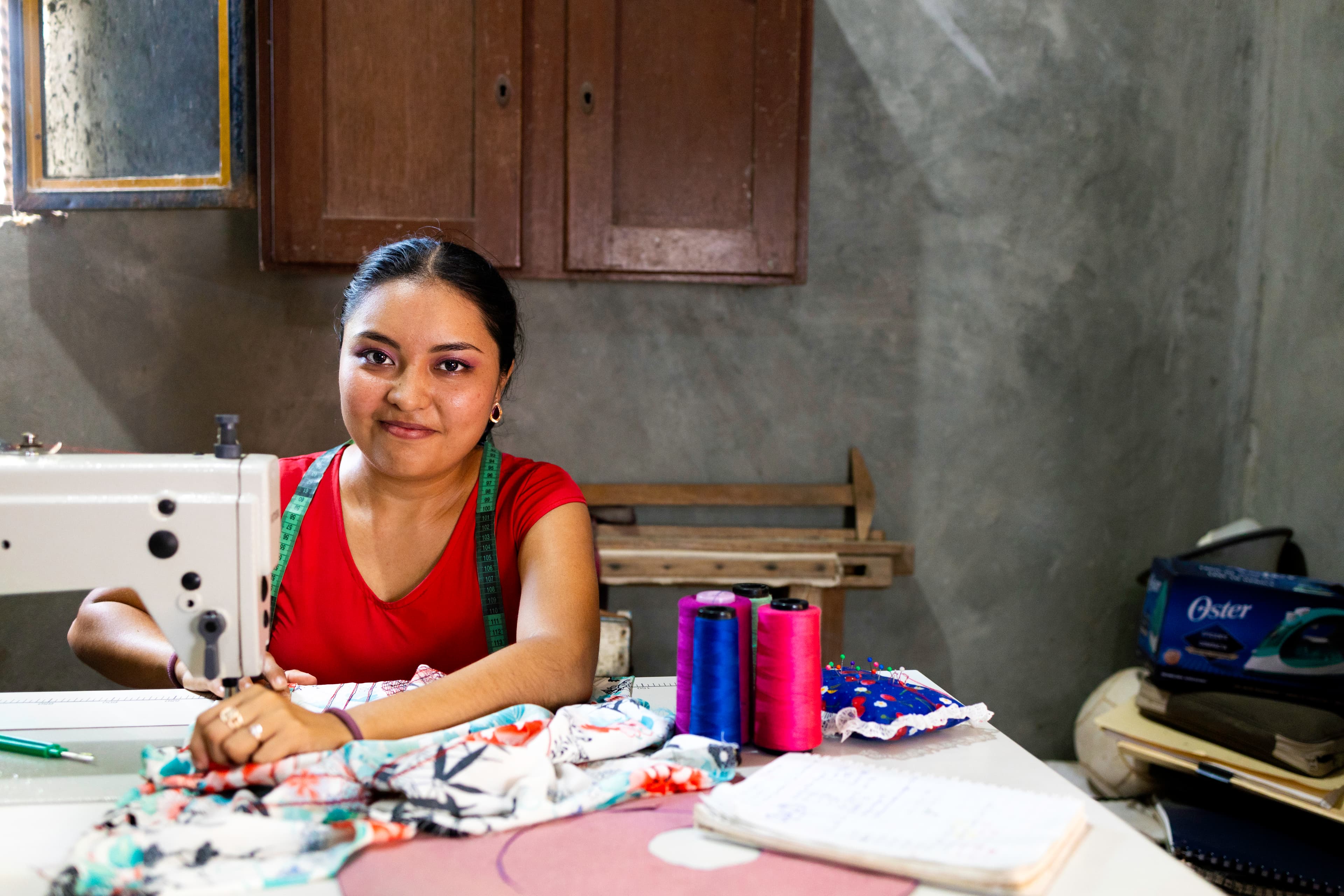 A young woman sits in front of a sewing machine while sewing and smiling.