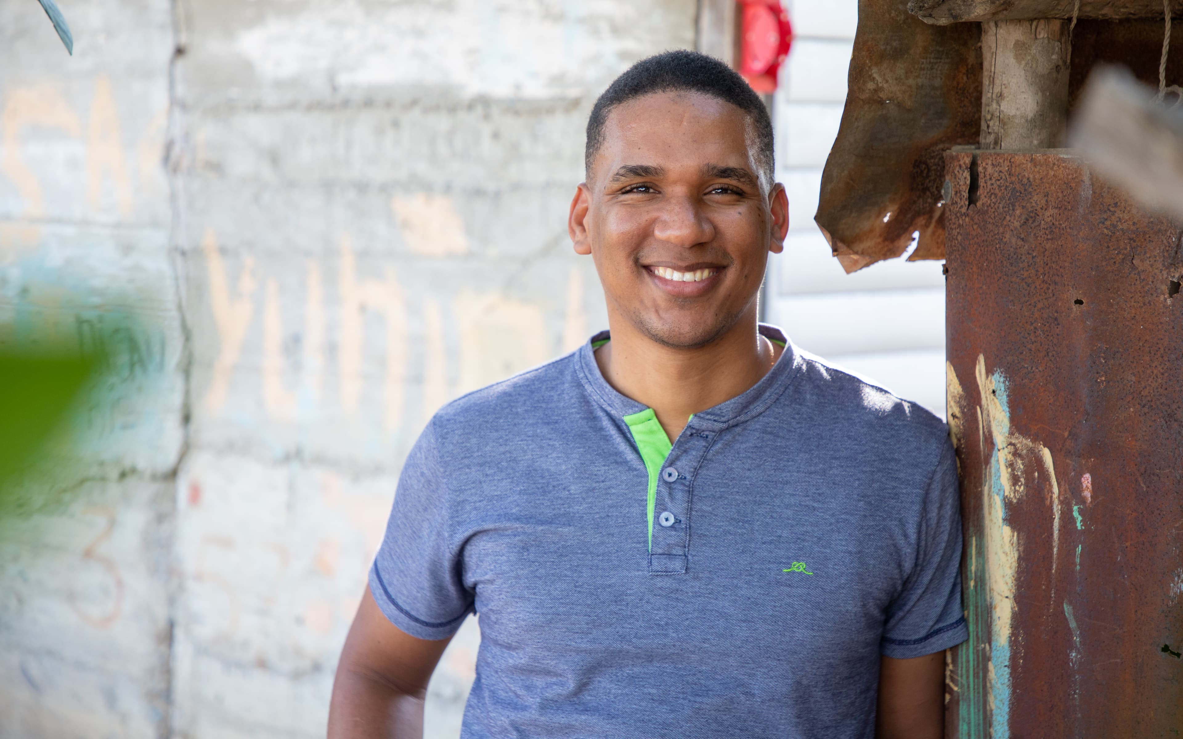 A man wearing a blue t-shirt leans against a building and smiles at the camera.