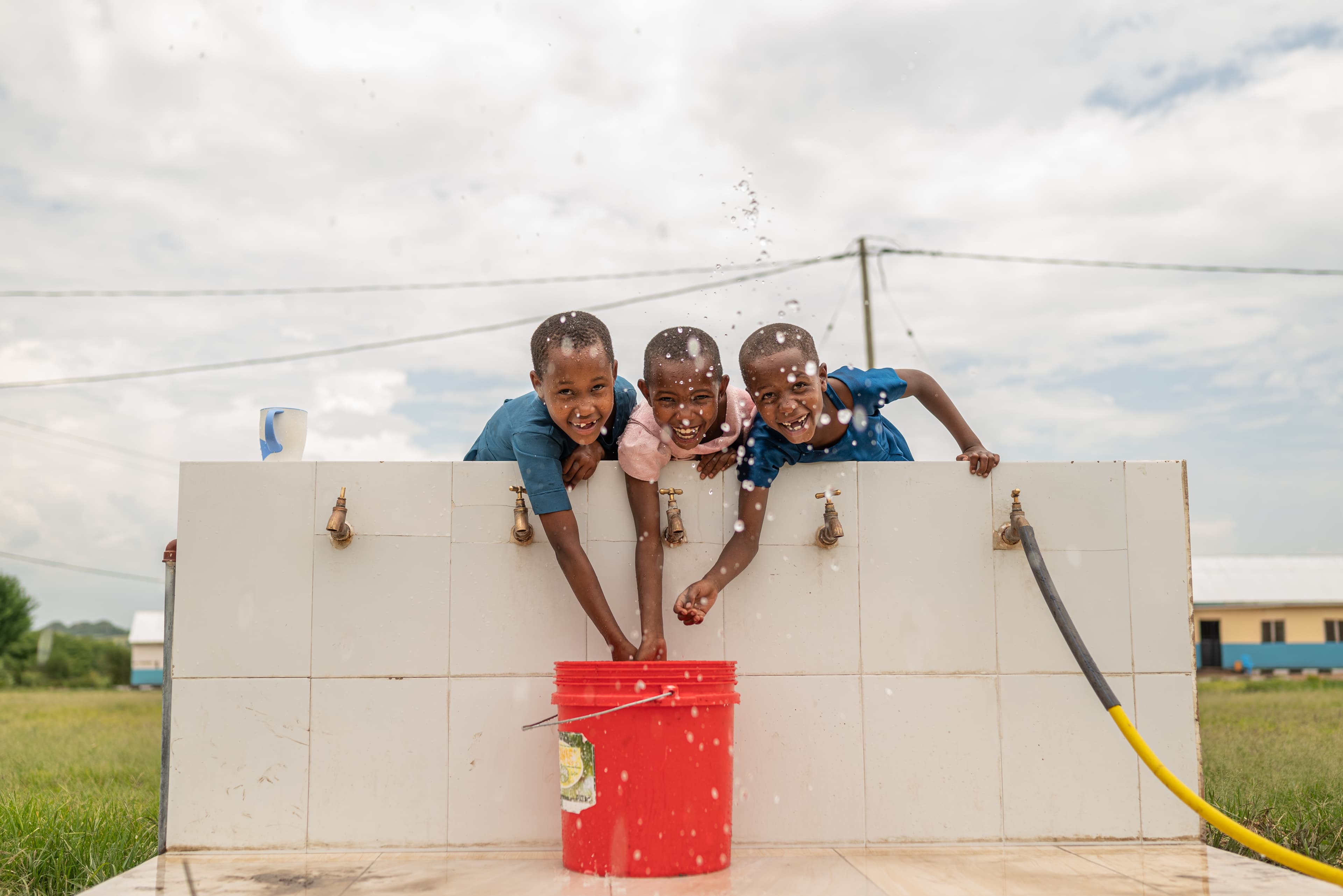 Three children lean over the side of a wall and splash in water faucets while smiling.
