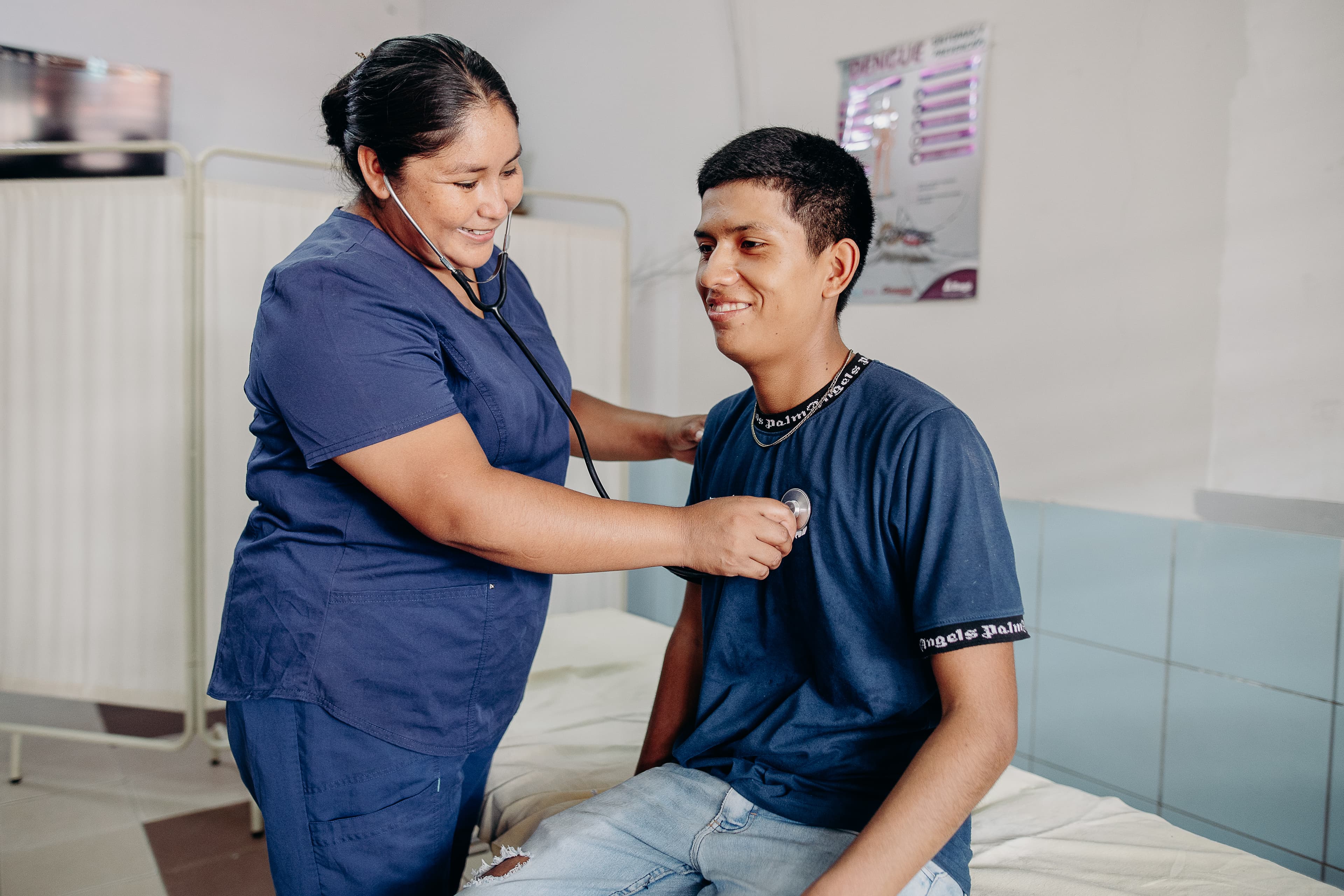A young man sits on a hospital bed smiling while a nurse checks his heart with a stethoscope.